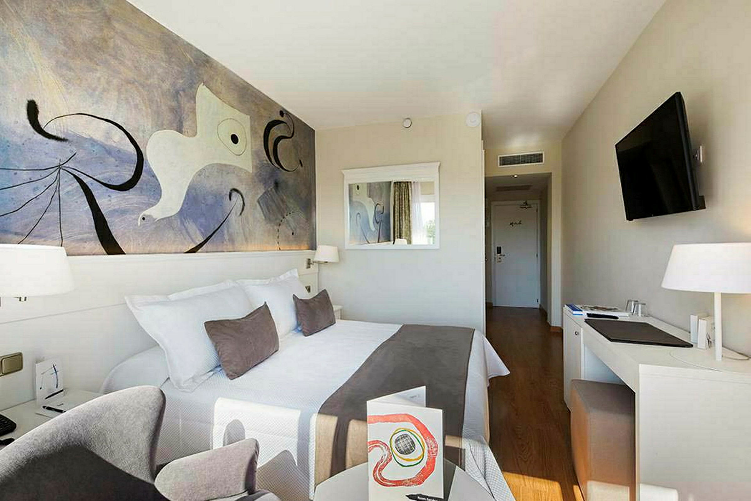 Ett hotellrum med en dubbelsäng, väggkonst, spegel, skrivbord och en TV. (AI-genererad text)
