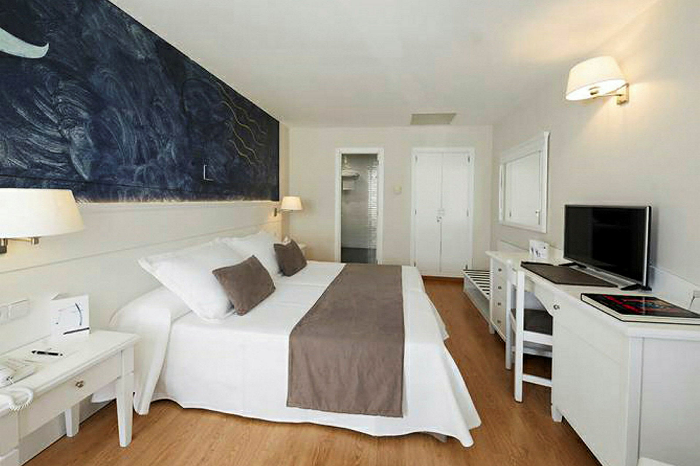Ett hotellrum med en dubbelsäng med vita och bruna sängkläder, en målning på väggen, och ett skrivbord med en tv. (AI-genererad text)