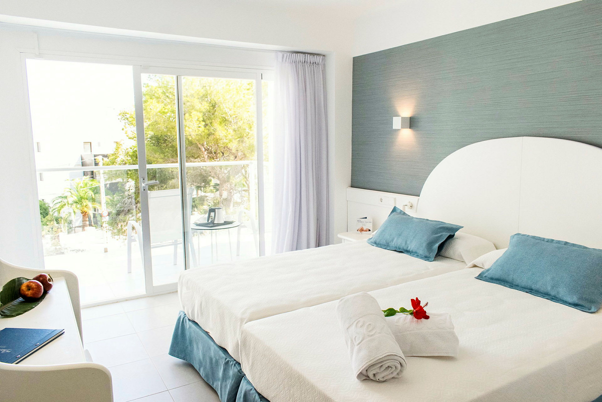 Hotellrum med bäddad säng och insläpp av dagsljus genom stora glasdörrar. (AI-genererad text)