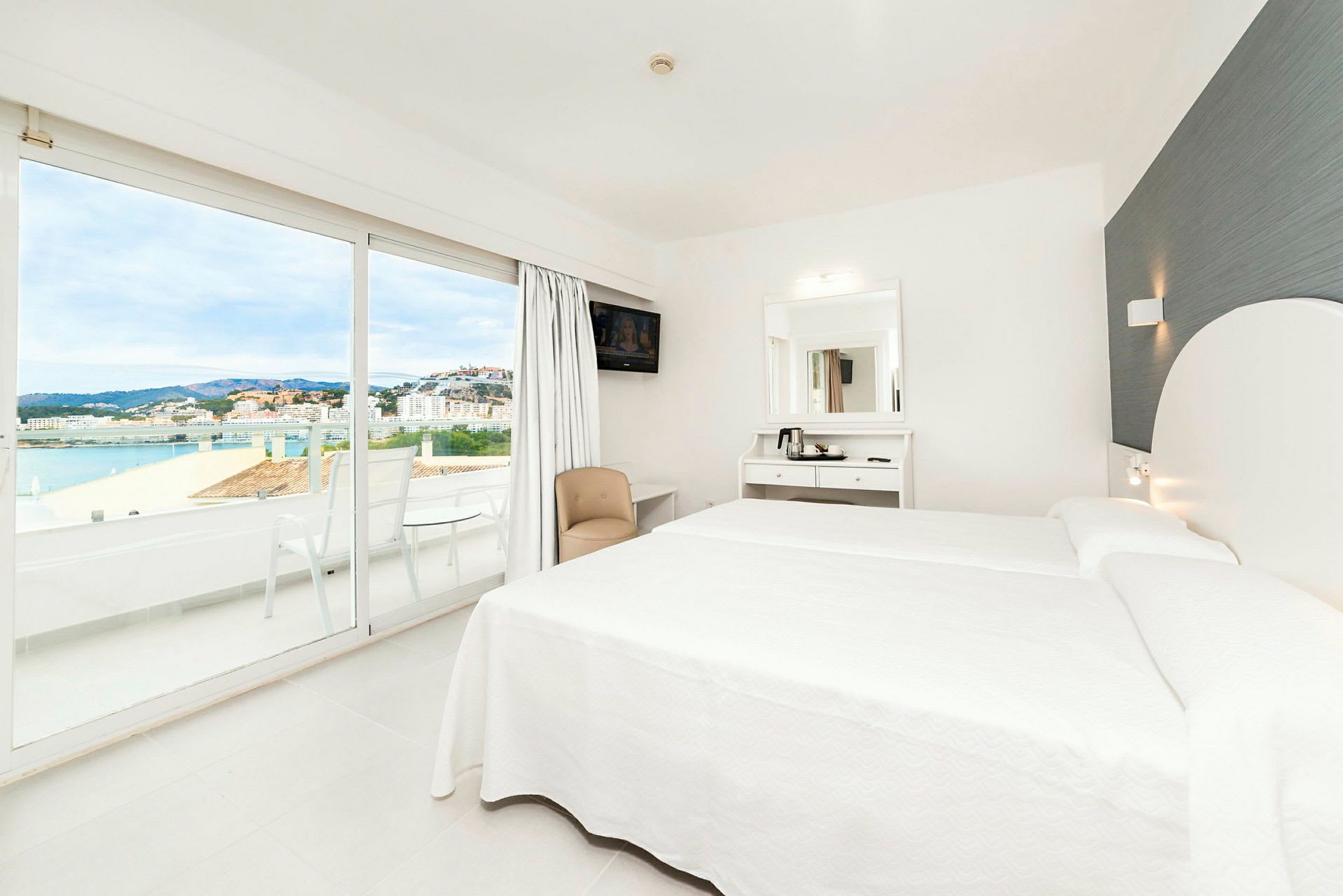 Ett modernt hotellrum med vita möbler, en dubbelsäng och stora glasdörrar som öppnar upp till en balkong med utsikt över en stad och havet. (AI-genererad text)