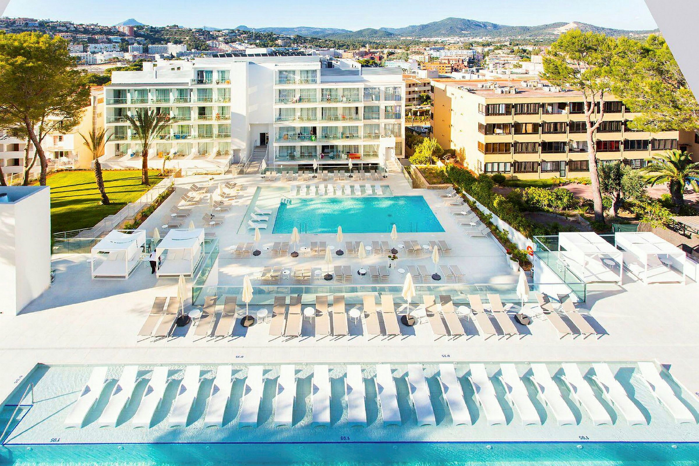 Vy över ett hotellområde med pooler, solsängar och parasoller, samt omgivande byggnader och grönska. (AI-genererad text)