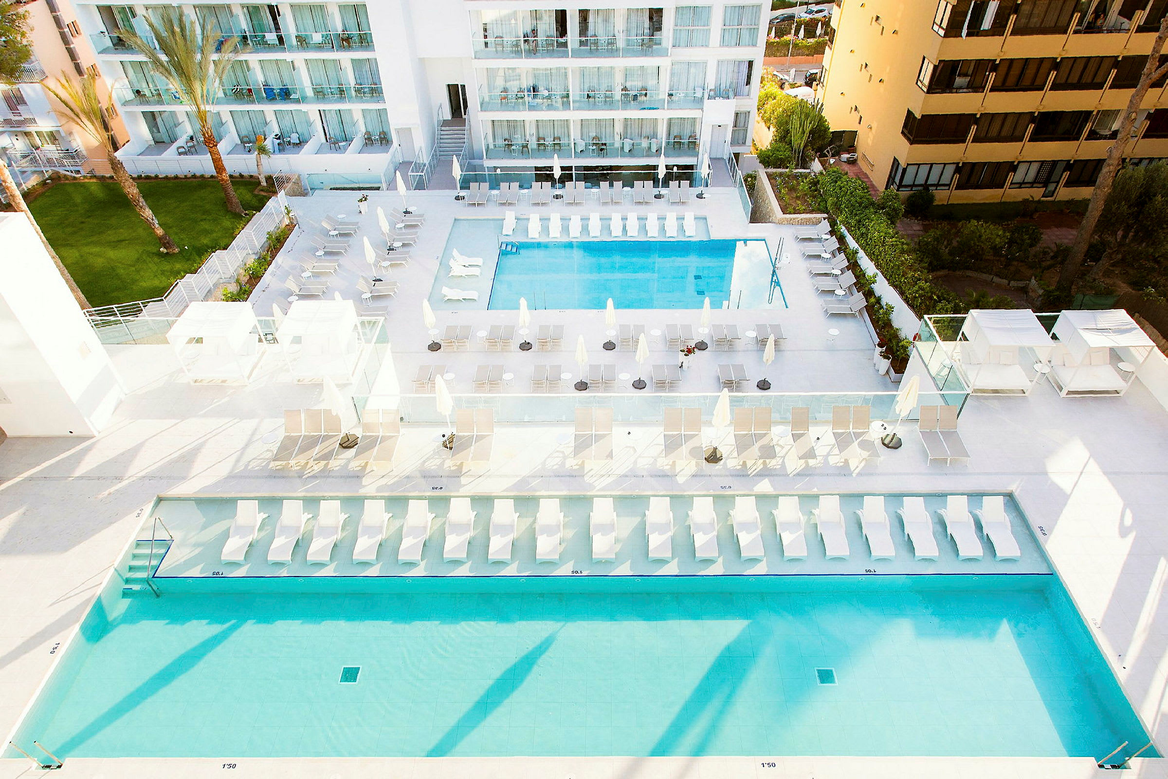 Bild föreställande en hotellpoolområde med två pooler, strandstolar och parasoller, omgiven av moderna byggnader. (AI-genererad text)