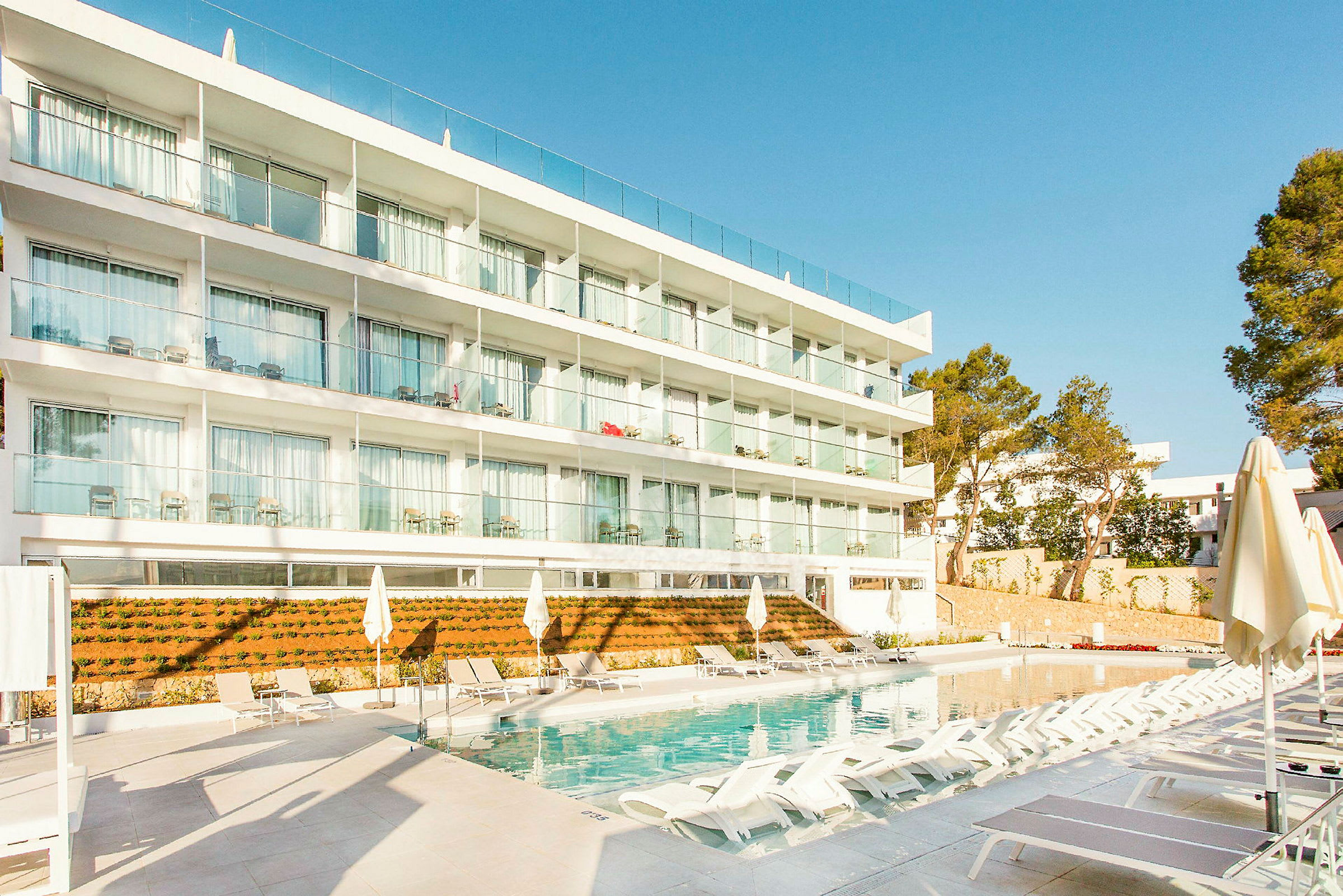 En hotellbyggnad med flera våningar och balkonger framför en swimmingpool omgiven av solstolar och parasoller. (AI-genererad text)