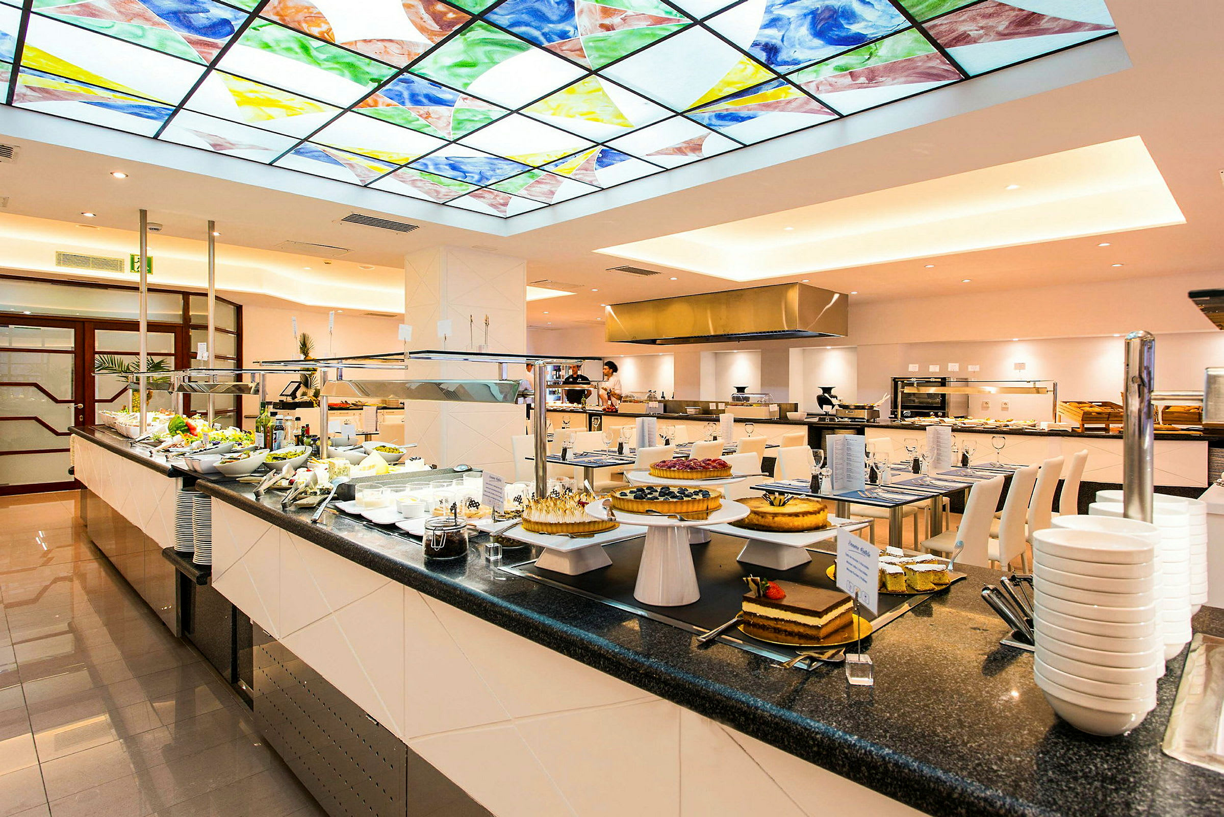 Bilden visar en matbuffé i en restaurang med ett färgglatt glastak ovanför. På buffén finns olika maträtter och desserter upplagda. (AI-genererad text)