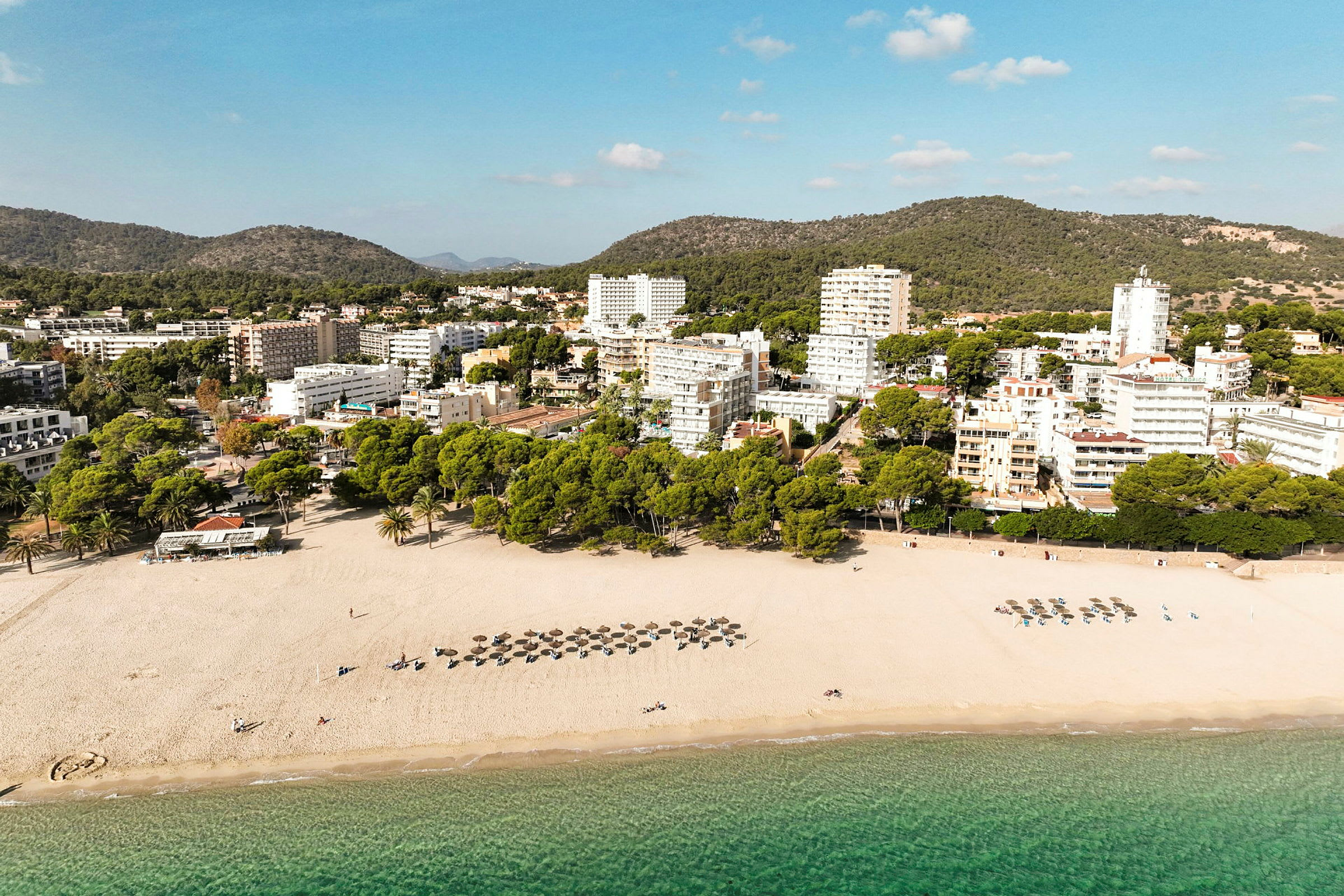 Strand med parasoller och stolar bredvid gröna träd och byggnader i bakgrunden. (AI-genererad text)