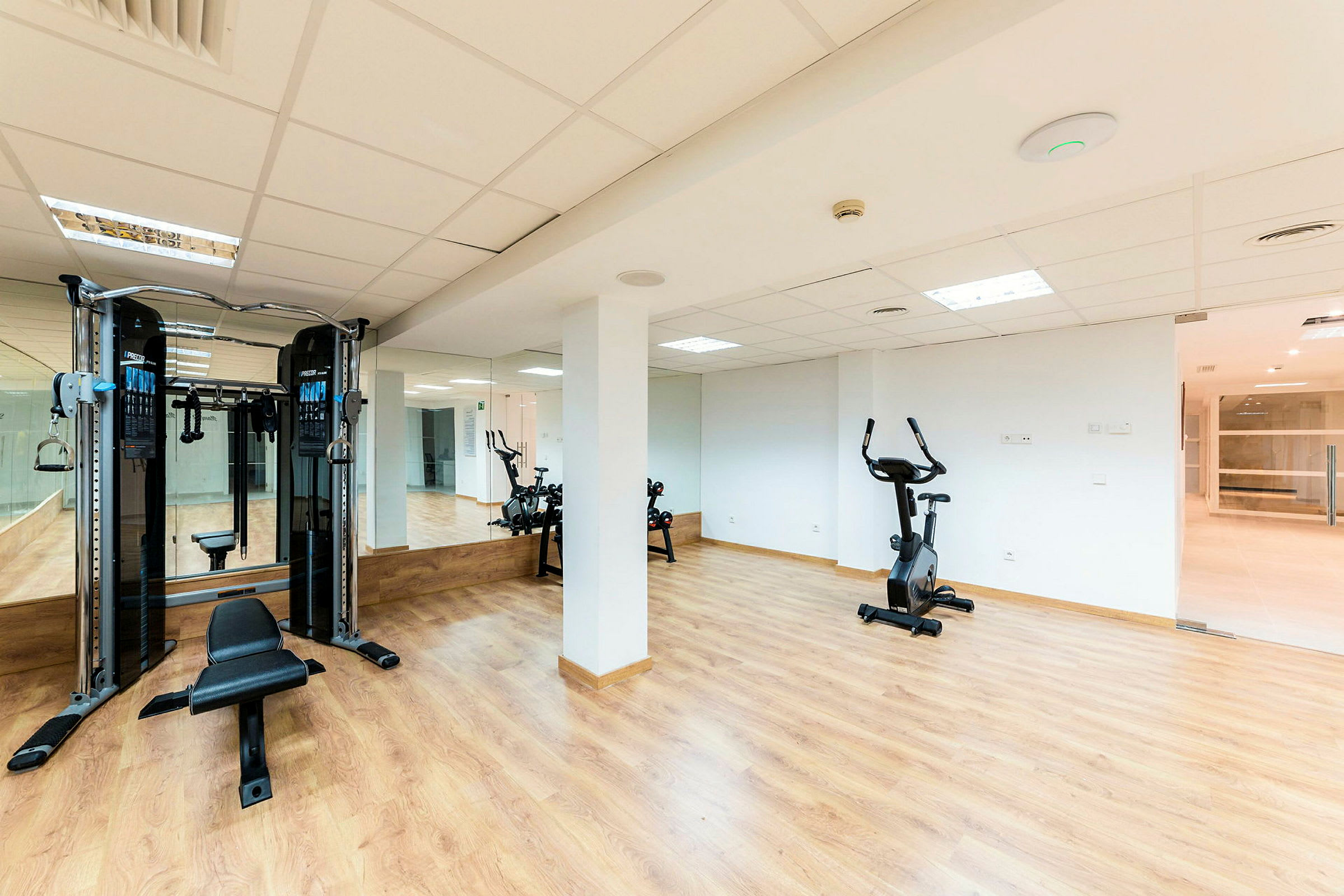 Ett ljust gymrum med träningsmaskiner, inklusive en kabelmaskin och en motionscykel, samt en väg med spegel och parkettgolv. (AI-genererad text)