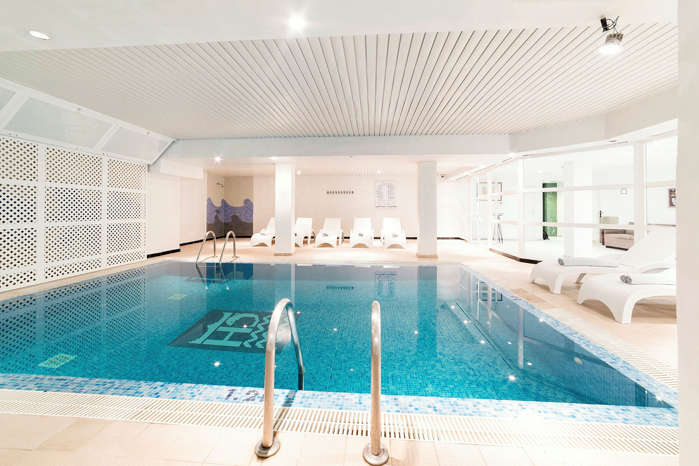 Bild av en inomhuspool med vita solstolar vid poolkanten och ett ljus och rent intryck. (AI-genererad text)