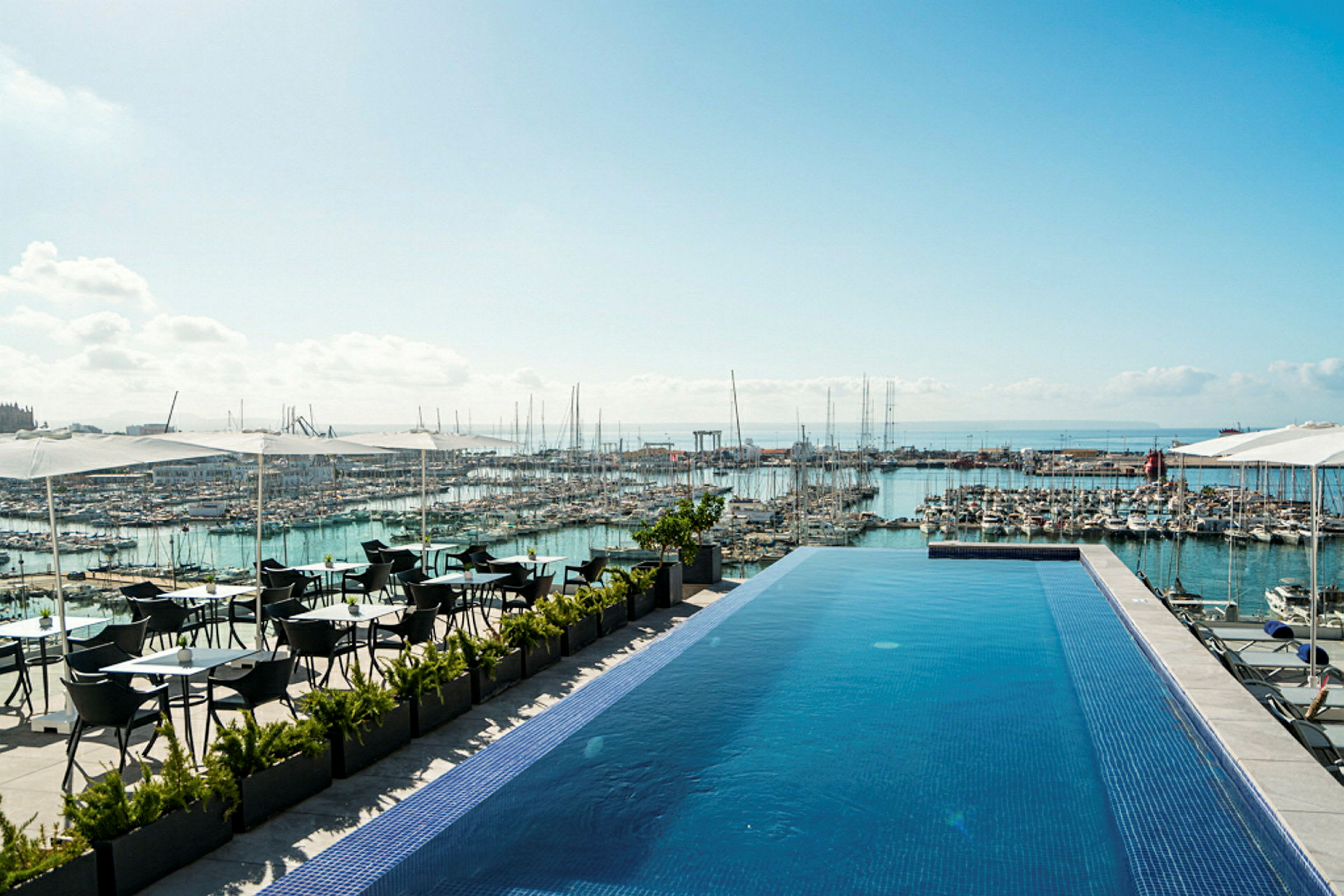 En infinitypool vid en terrass med utsikt över en marina full av båtar och havet i bakgrunden. Det finns sittplatser under parasoller intill poolen. (AI-genererad text)