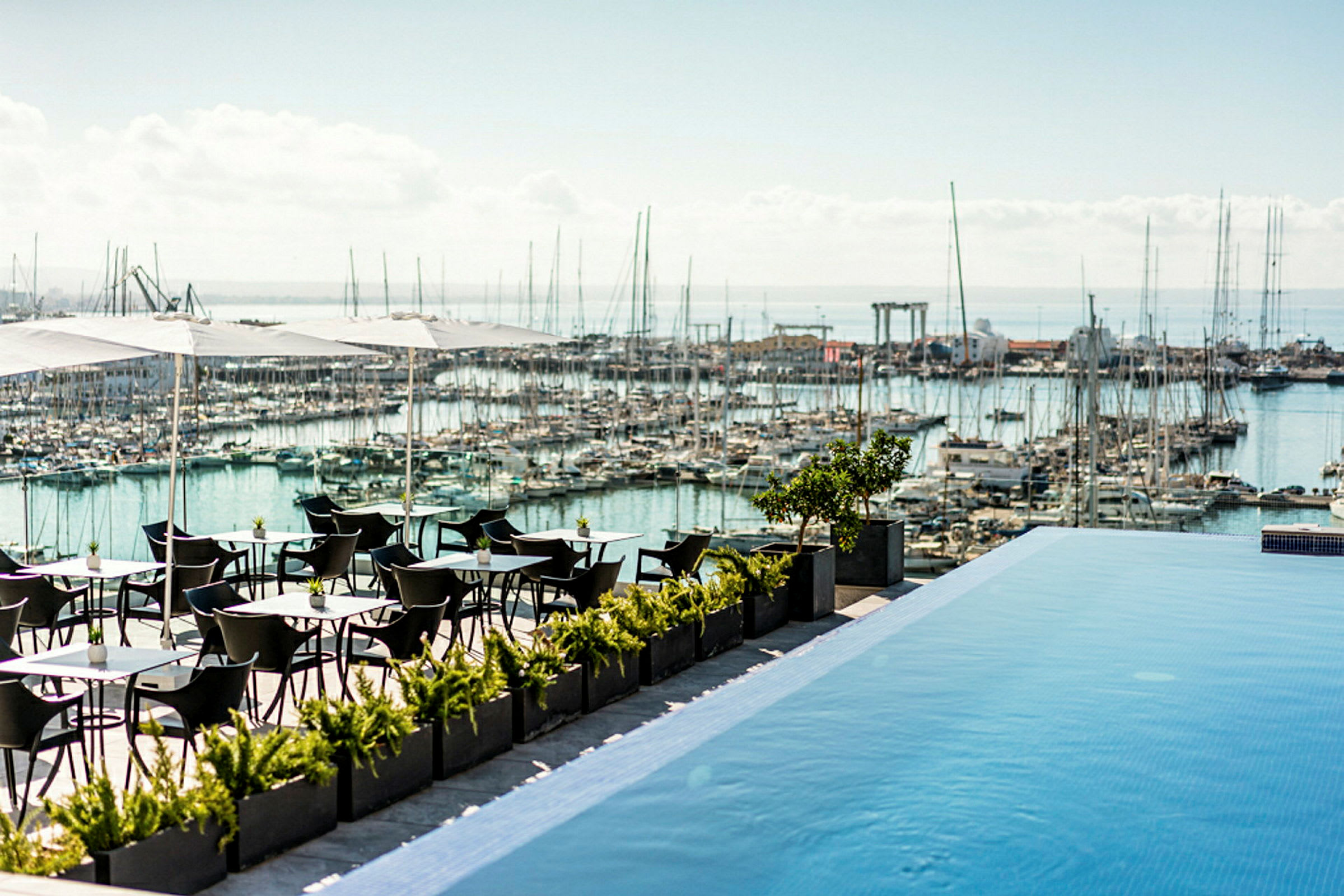 En utomhusterrass med bord och stolar under parasoller, bredvid en infinitypool med utsikt över en marina fylld med båtar. (AI-genererad text)