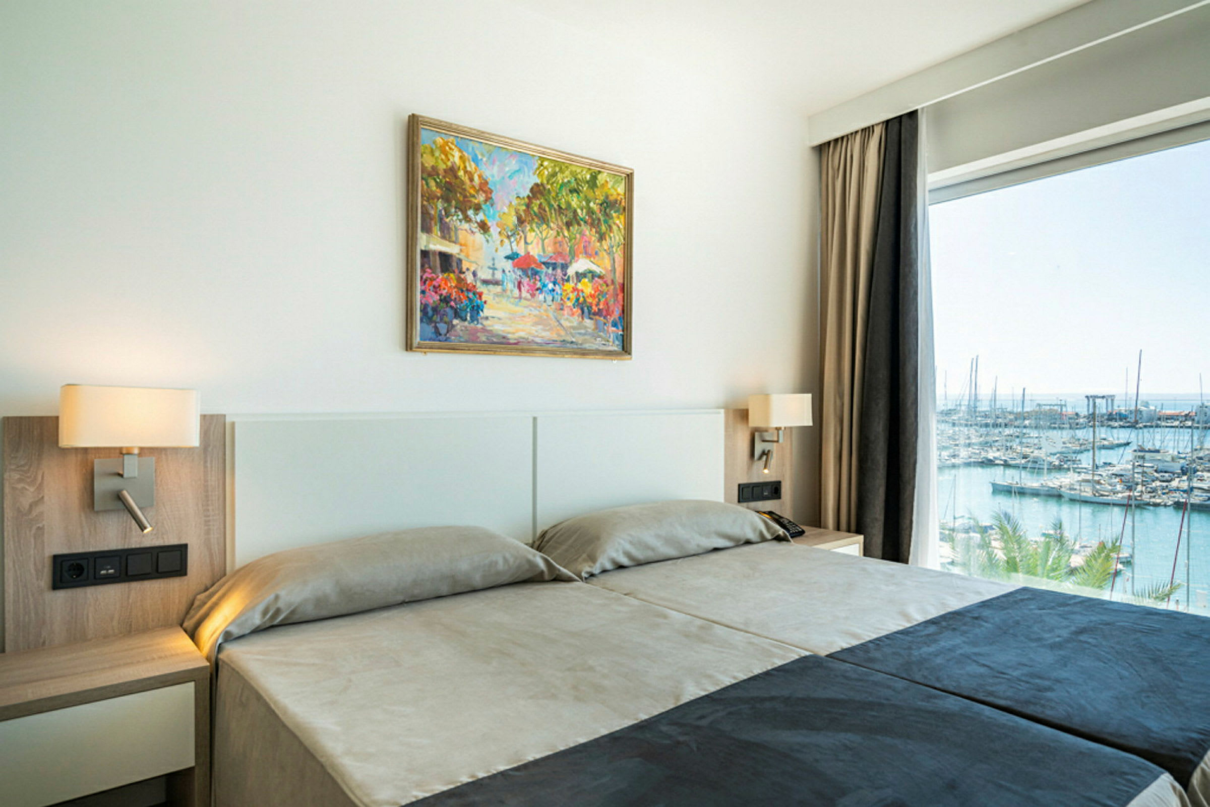 Ett hotellrum med två sängar, vägglampor, en färgglad målning på väggen och en stor fönsteröppning med utsikt över en marina. (AI-genererad text)