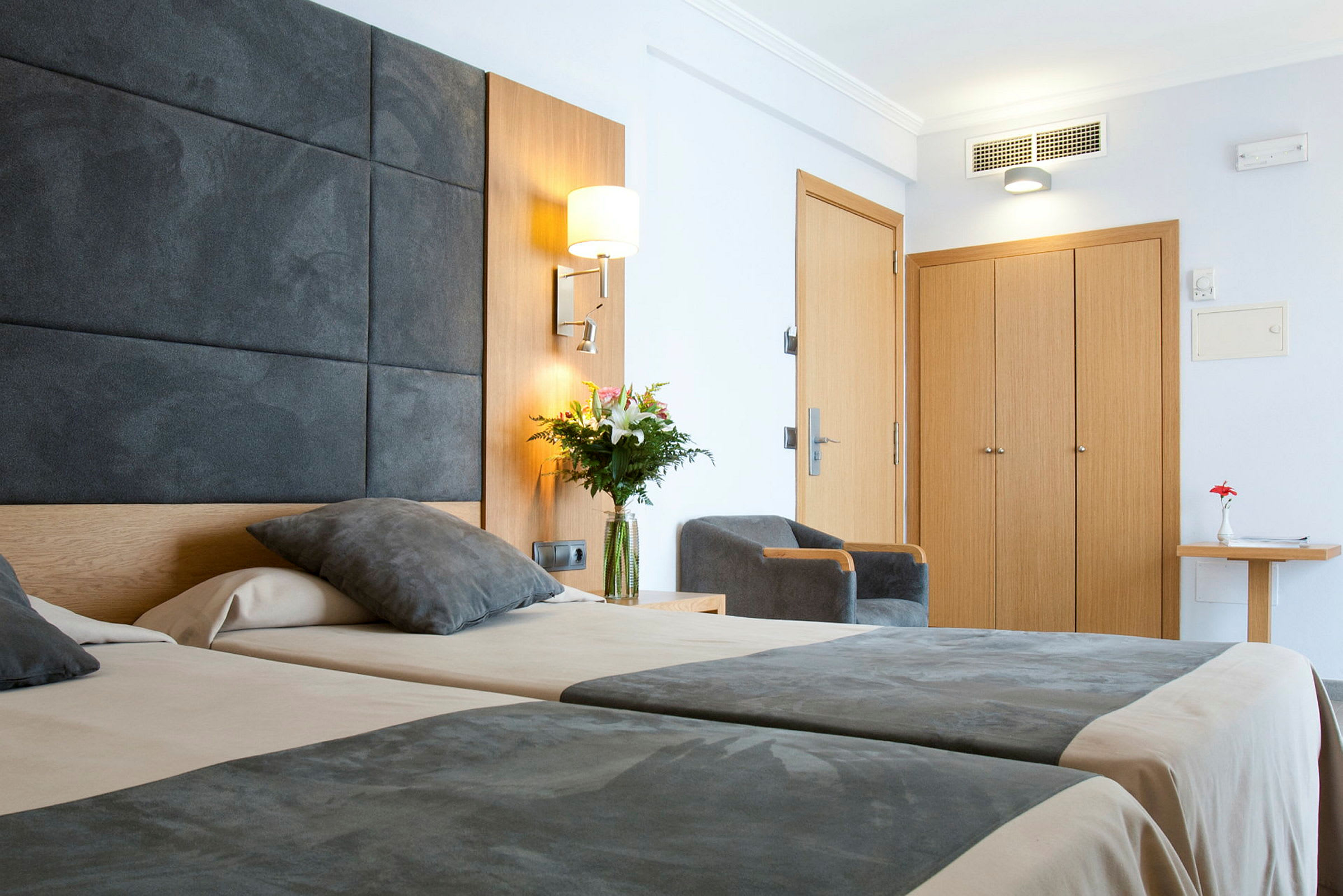 Bild av ett modernt hotellrum med två sängar, gråa överkast, en stol, en garderob i trä och en blomvas på ett bord. (AI-genererad text)