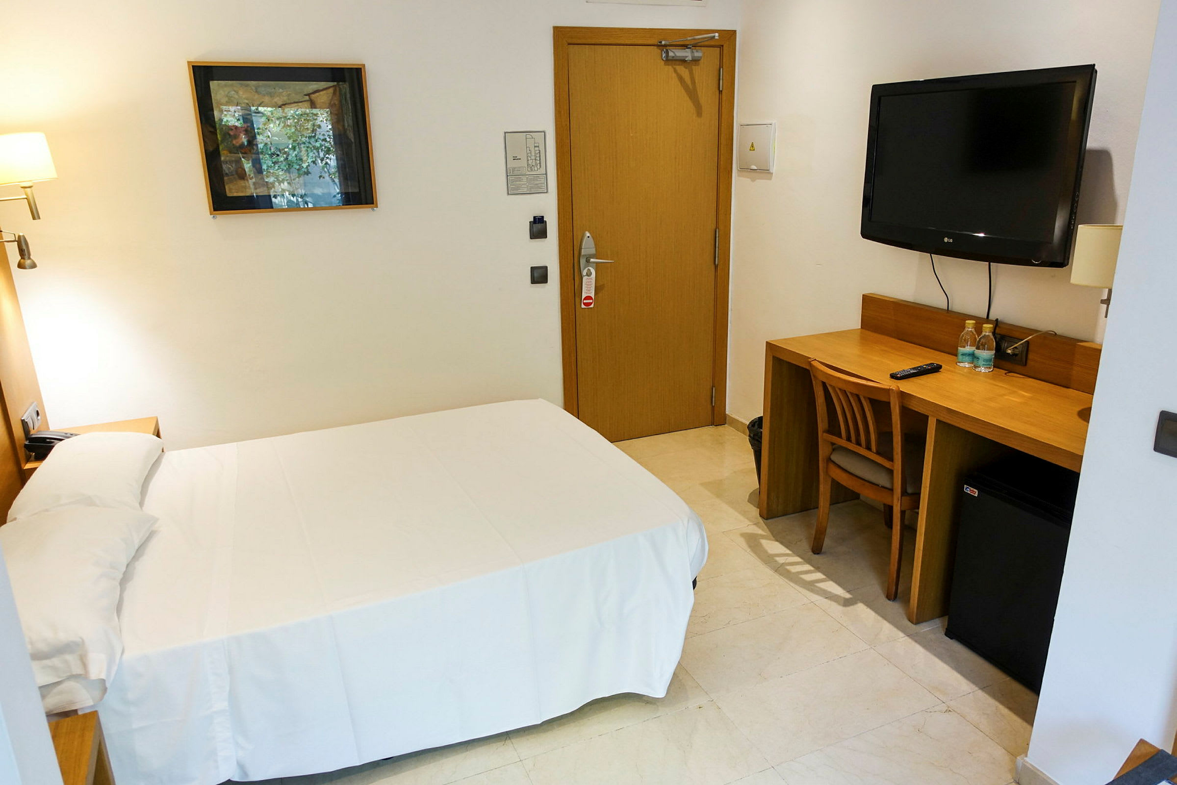 Ett enkelt hotellrum med en säng, skrivbord och stol. På väggen finns en TV och ett konstverk. (AI-genererad text)