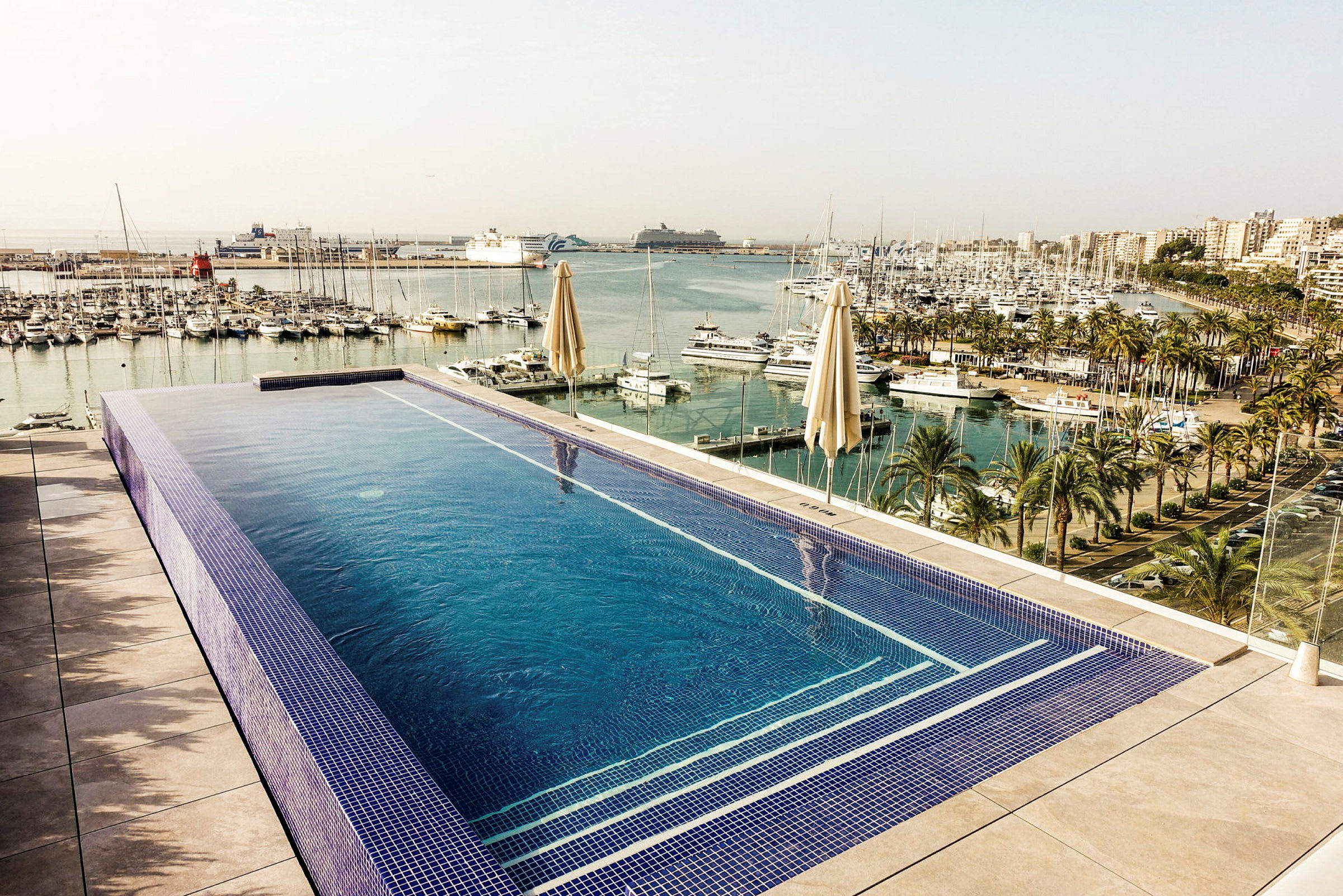 Bild på en infinitypool med utsikt över en småbåtshamn med palmer och flervåningsbyggnader i bakgrunden. (AI-genererad text)