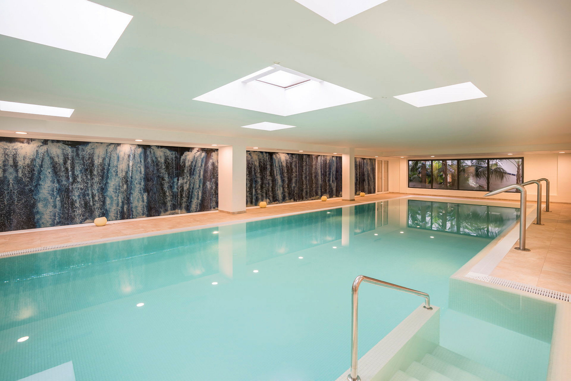 En inomhuspool med en vattenfallsväggdekoration och ljusa takfönster. (AI-genererad text)