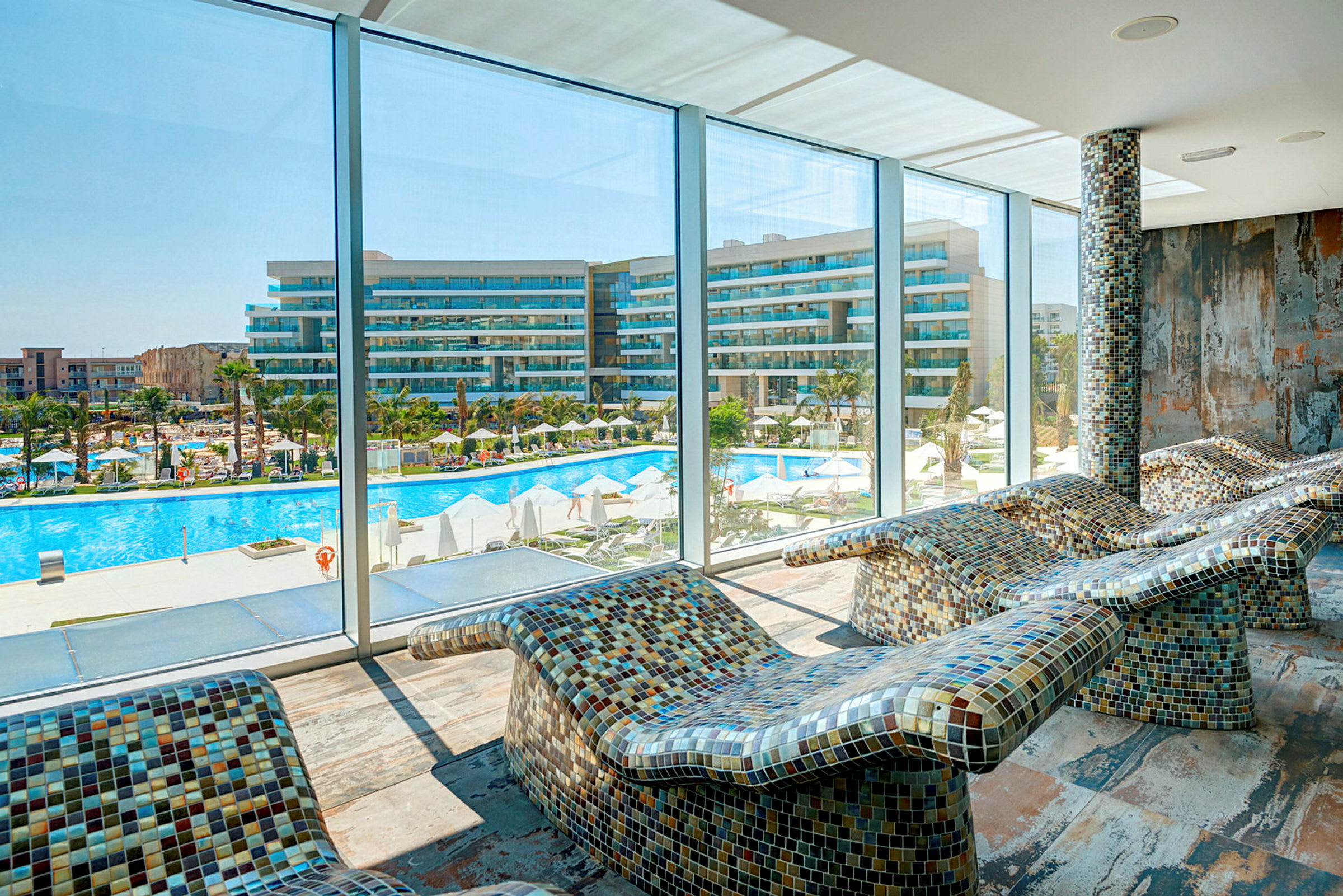 Utsikt från ett rum med mosaikbeklädda vilstolar mot en pool och ett hotell. (AI-genererad text)