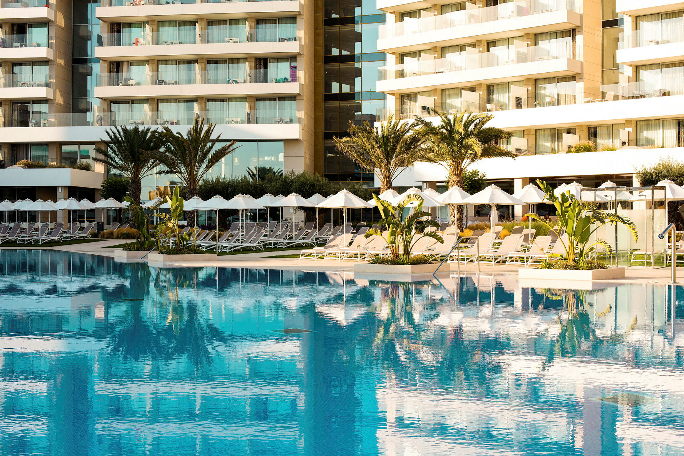 En stor pool framför en modern hotellbyggnad med solstolar och parasoller. (AI-genererad text)