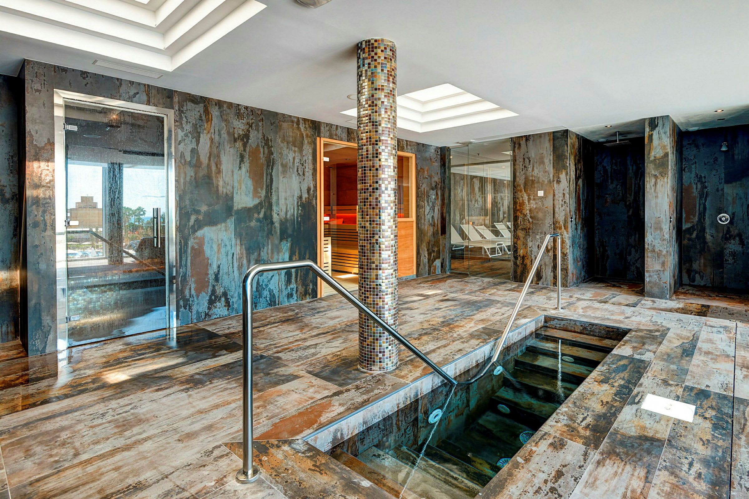 Interiör av ett spa med pool, mosaikbeströdd pelare och sittområden. (AI-genererad text)