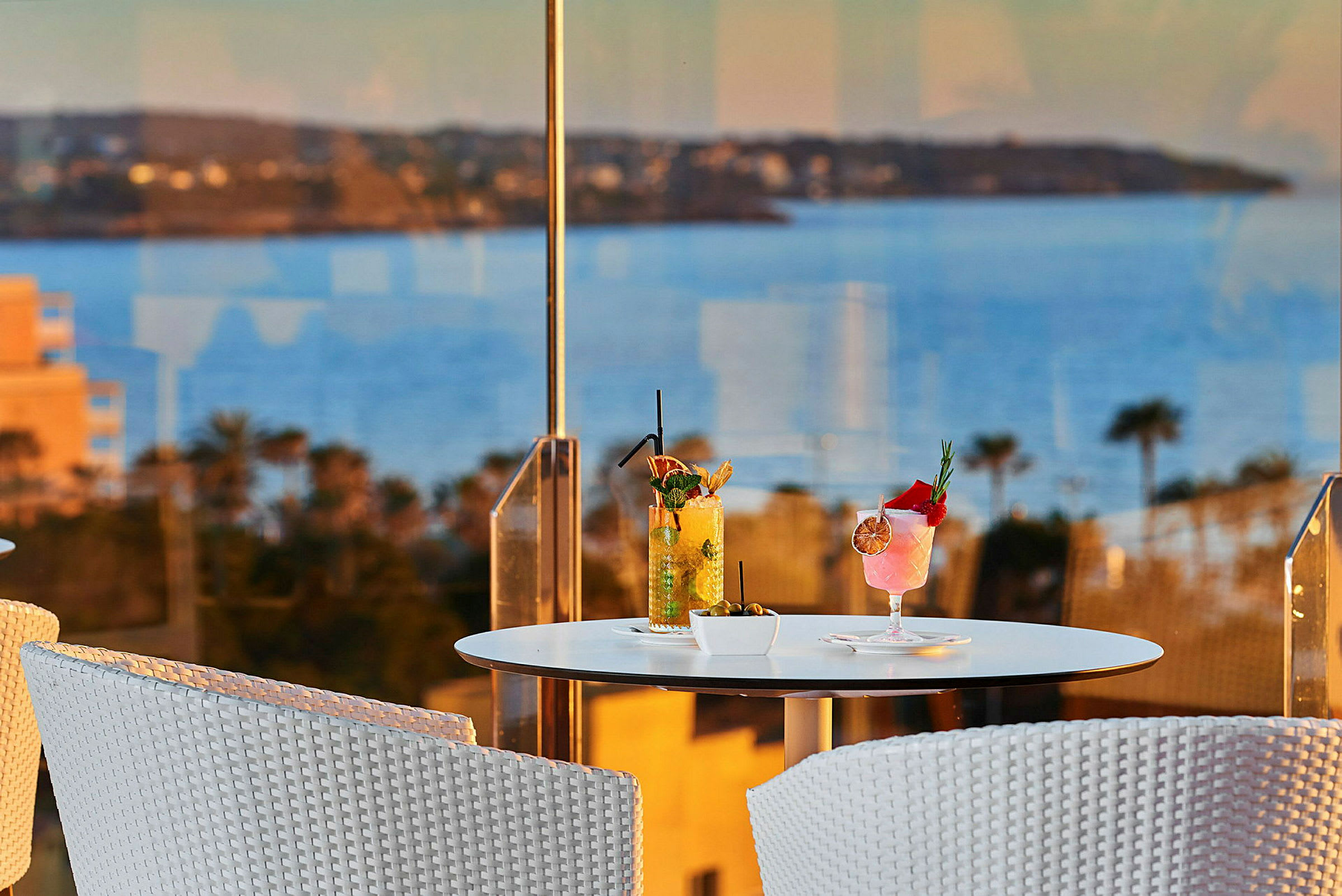 En vit sittgrupp med ett bord där två cocktails står, med havsutsikt i bakgrunden. (AI-genererad text)