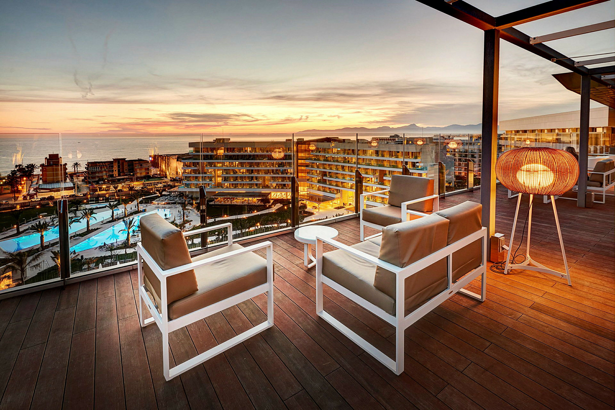 Utsikt över solnedgången från en terrass med sittmöbler och en golvlampa, med hotell och poolområde i bakgrunden. (AI-genererad text)