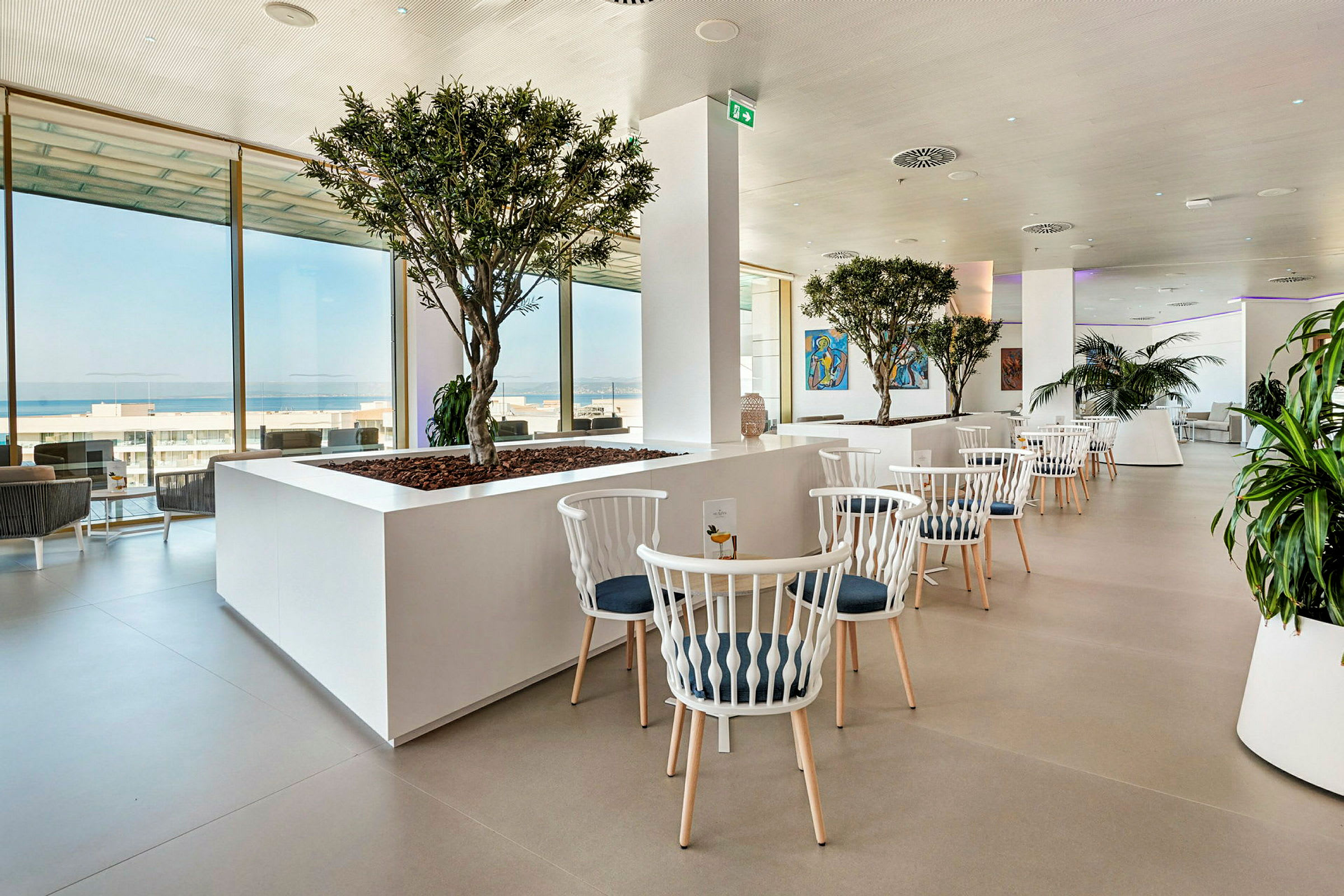 Elegant restaurang med vita möbler, gröna växter och stora fönster med utsikt. (AI-genererad text)