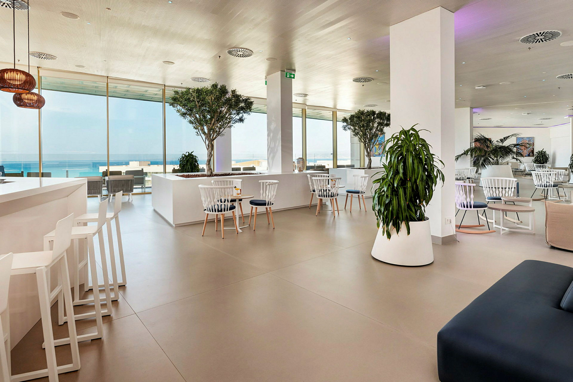 Modern lounge med vita möbler och gröna växter, havsutsikt genom stora fönster. (AI-genererad text)