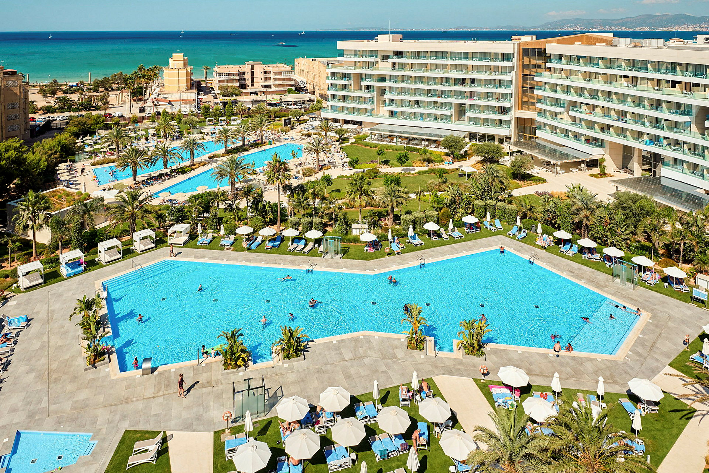 Vy över en semesterresort med pooler, solstolar och omgivande hotellbyggnader, samt havet i bakgrunden. (AI-genererad text)