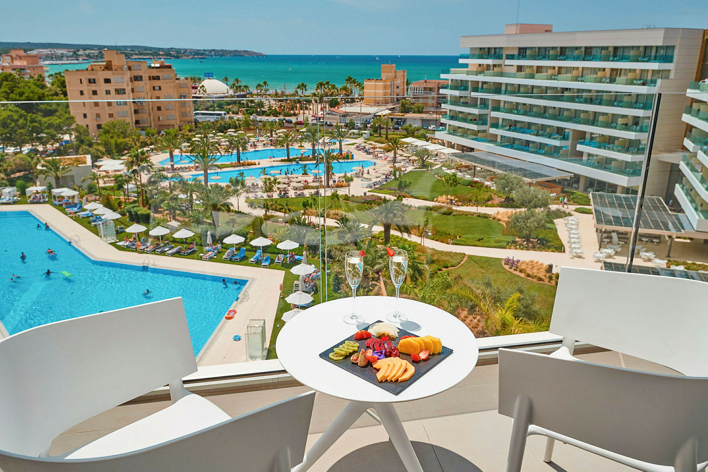 Utsikt från en balkong mot en hotellpool och havet, med en fruktbricka och två glas på ett bord. (AI-genererad text)
