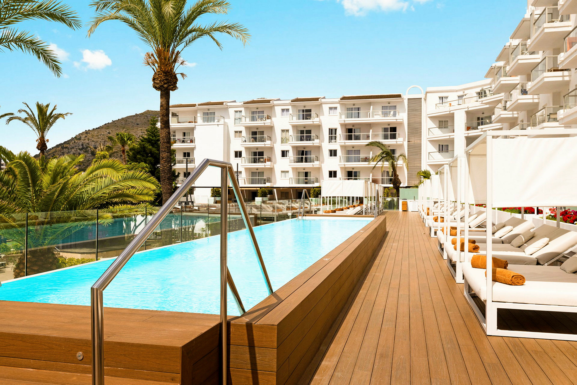 En pool omgiven av solstolar och vita hotellbyggnader, med palmer i bakgrunden. (AI-genererad text)