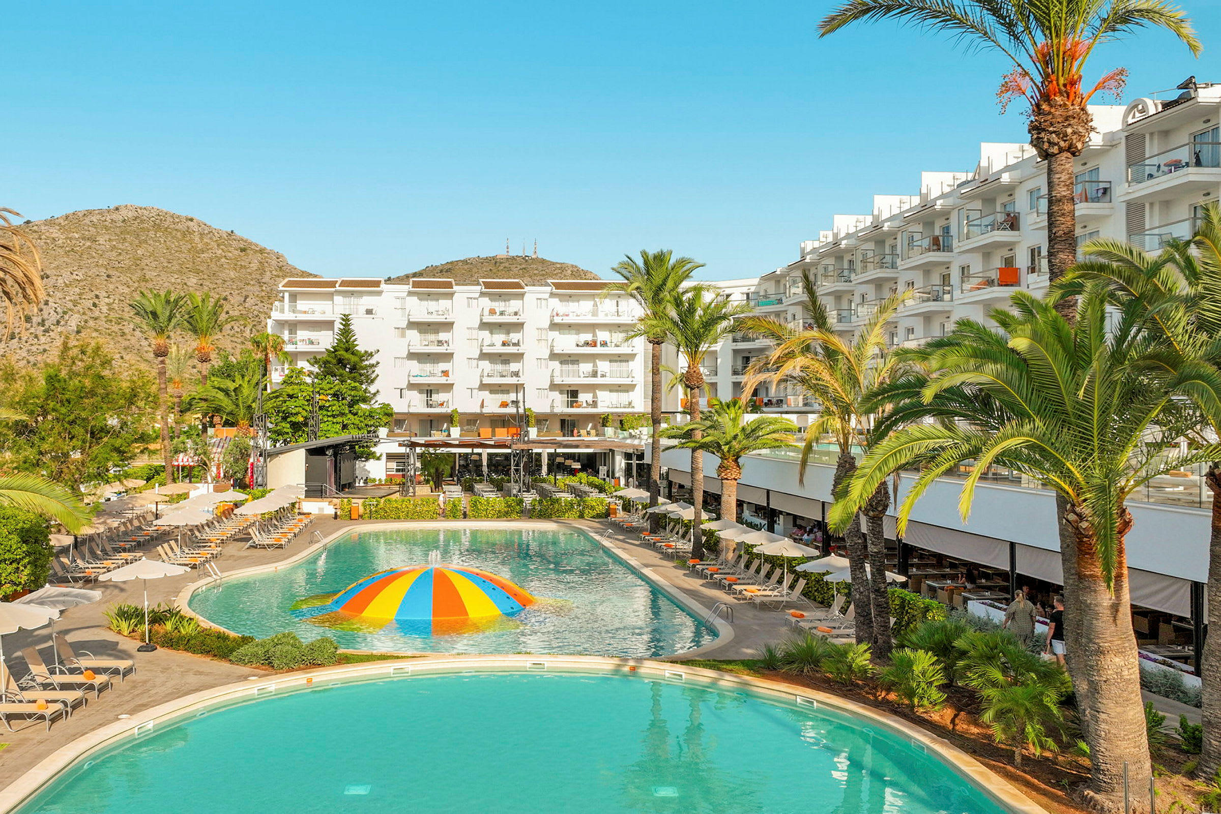 Ett hotell med vita byggnader, solstolar och en pool med en färgglad vattensprutande kupol i mitten. (AI-genererad text)