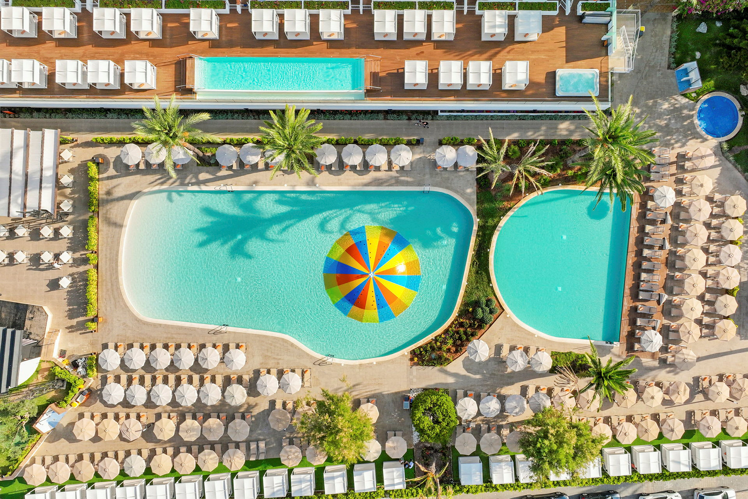 Flygfoto av en hotellpool med parasoller och solstolar runt omkring, samt en färgglad parasoll i vattnet. (AI-genererad text)
