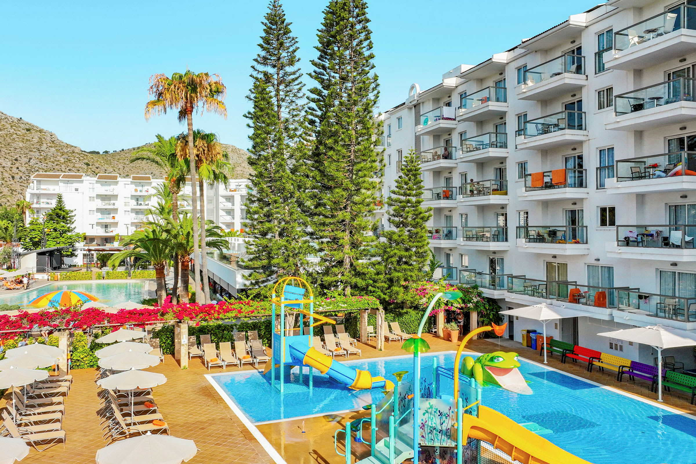 Vy över hotellpoolområde med vattenrutschkana, solstolar och omgivande byggnader. (AI-genererad text)