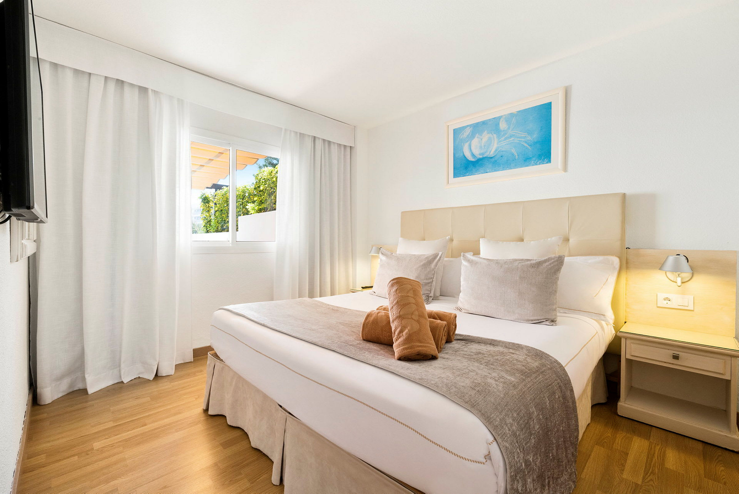 Ett hotellrum med en dubbelsäng, bäddad med prydnadskuddar och handdukar ovanpå, mot en bakgrund av ett fönster med vita gardiner. (AI-genererad text)