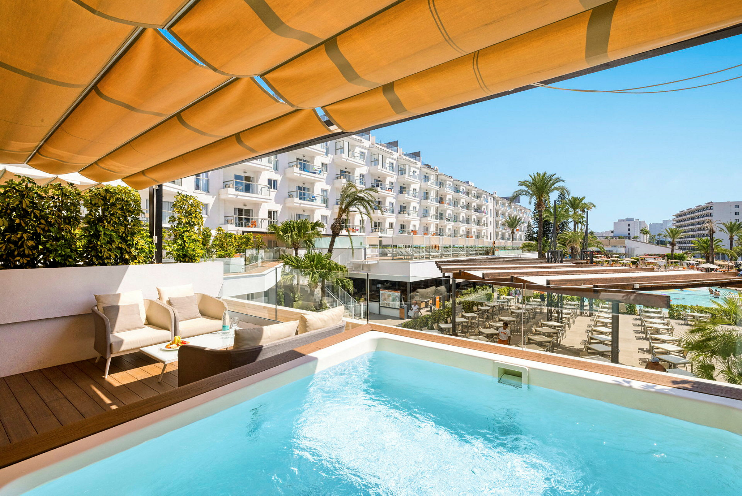 Terrass med pool, sittgrupp och utsikt över hotellområde med palmer och byggnader. (AI-genererad text)