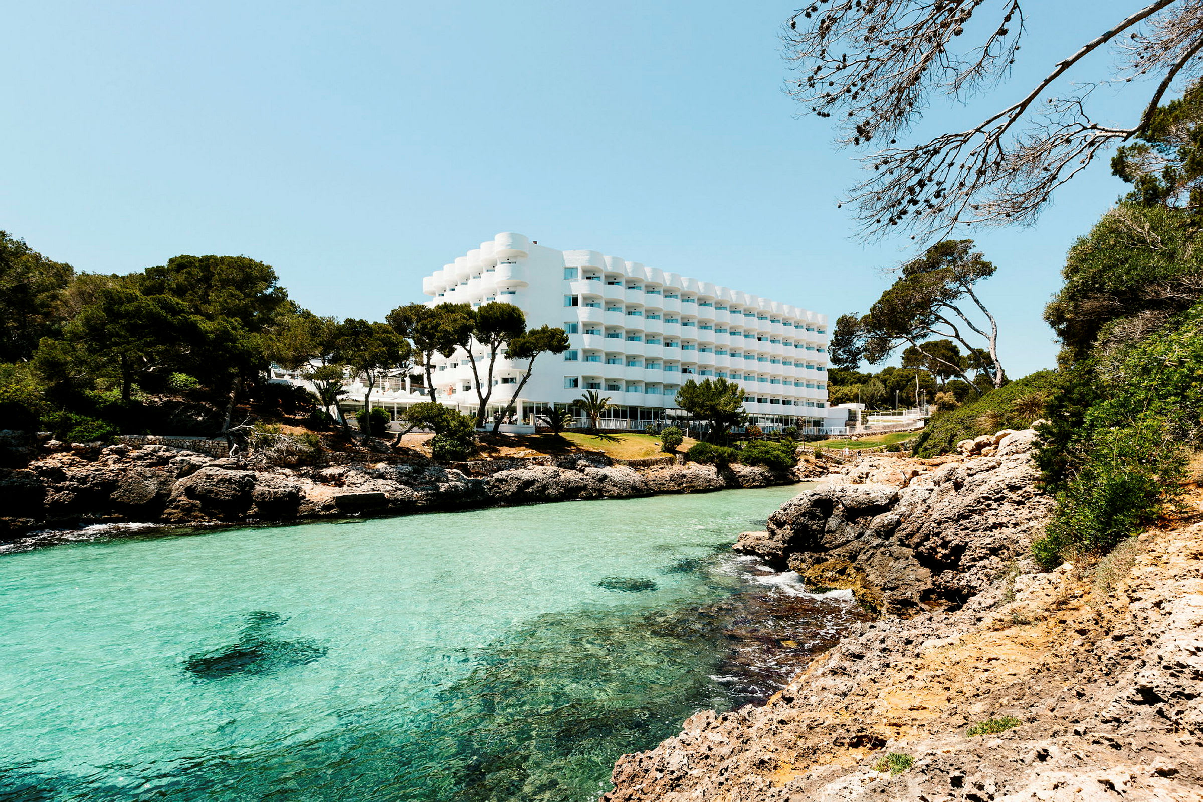 Hotellbyggnad med vit fasad, belägen vid en klippig kust med klart turkost vatten och omgivande grönska. (AI-genererad text)