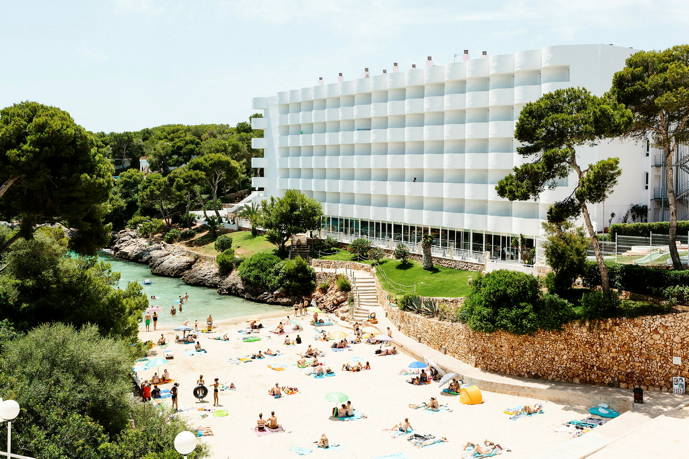 Strand med badande människor, omgiven av grönska och ett vitt hotell. (AI-genererad text)