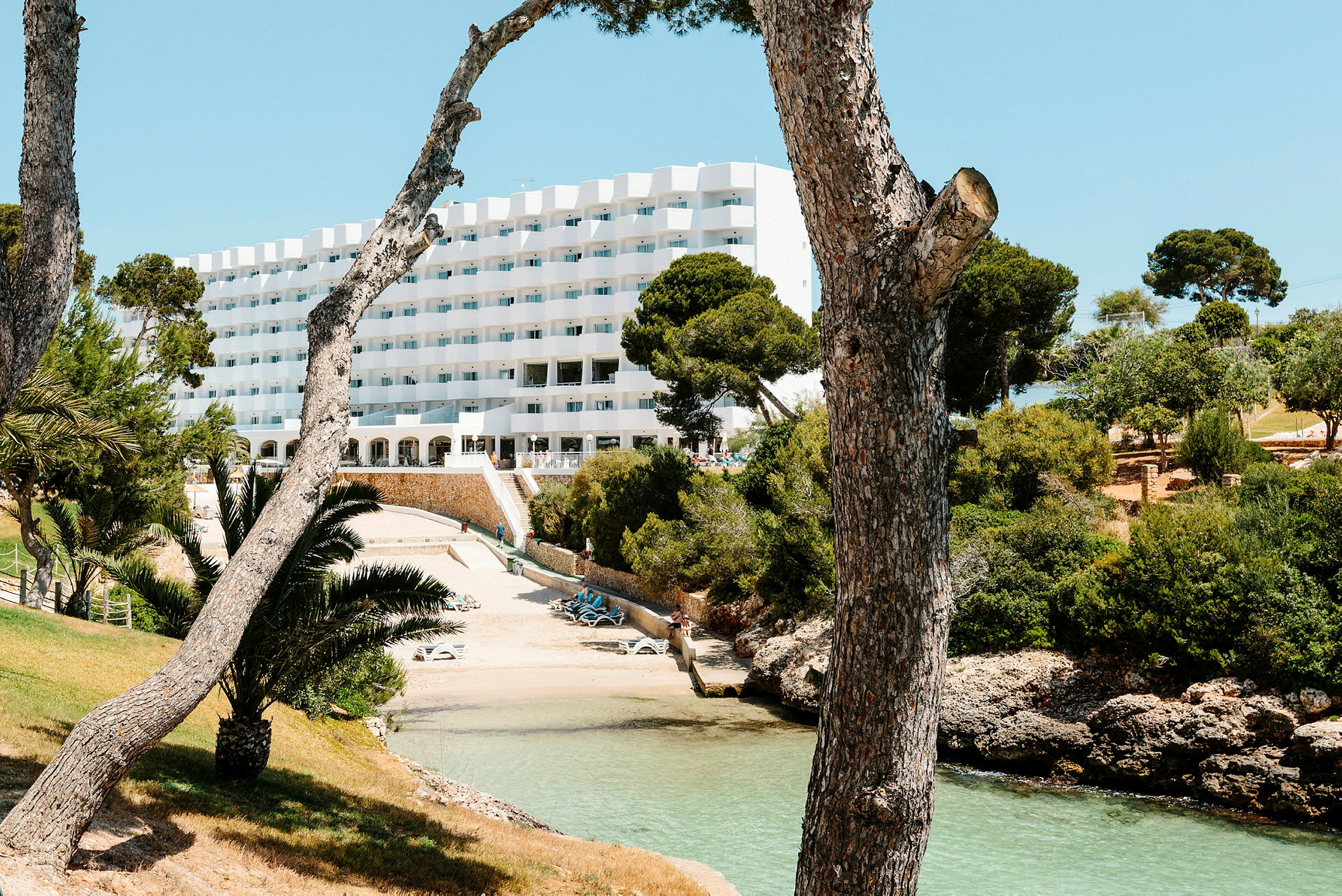 Hotellbyggnad belägen vid en liten strand, omgiven av träd och grönska. (AI-genererad text)