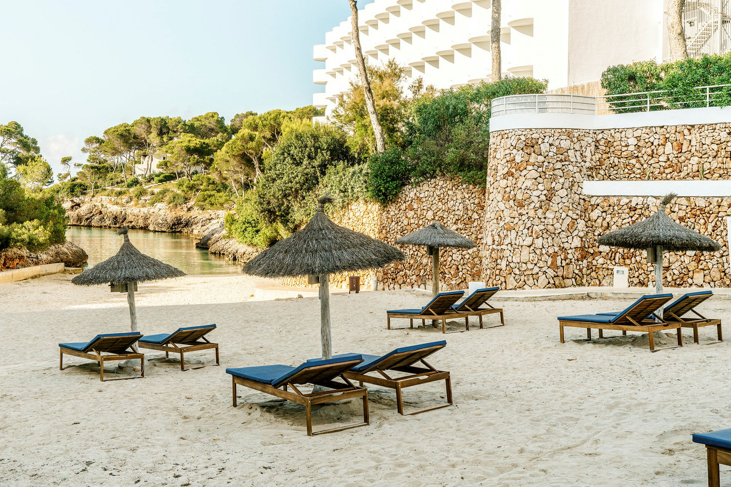 Solstolar och parasoller på en sandstrand, med stenmur och grönska i bakgrunden. (AI-genererad text)