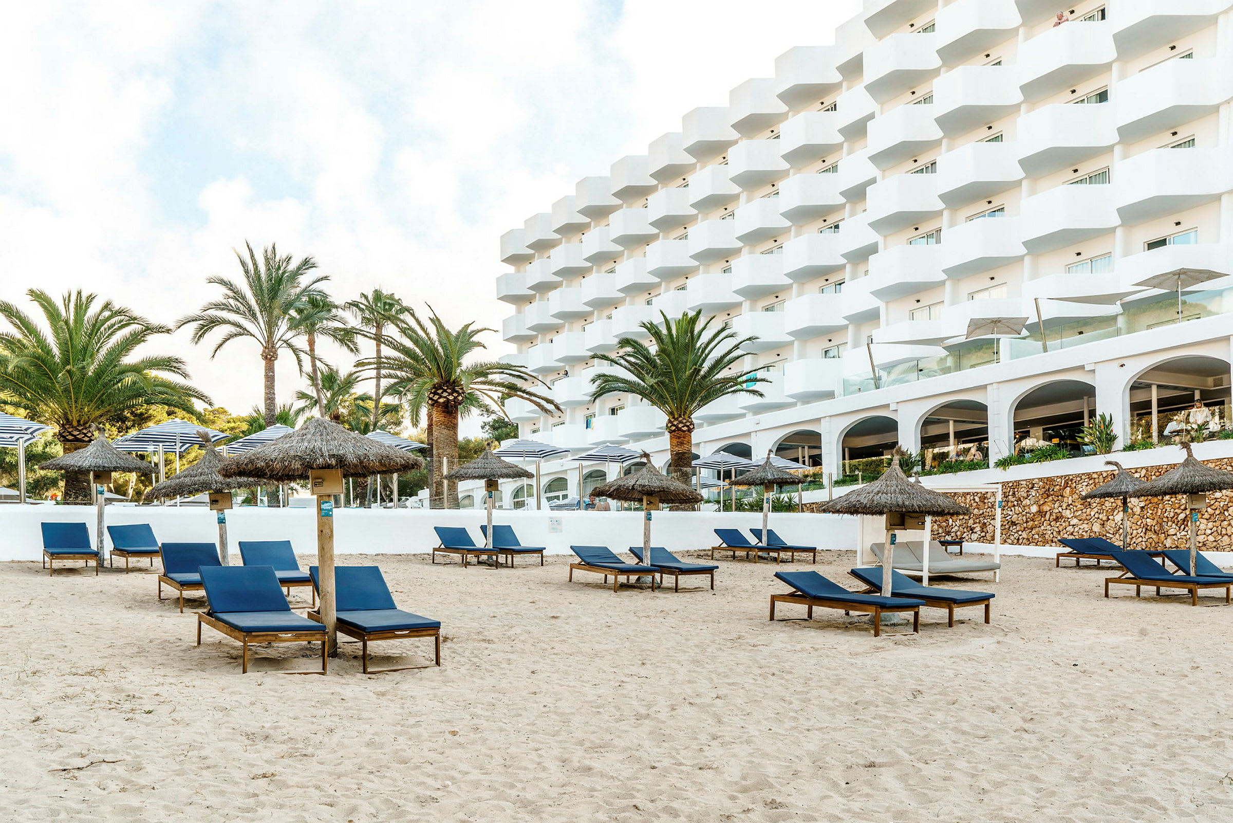 Strand med solstolar och parasoller framför en vit byggnad. (AI-genererad text)