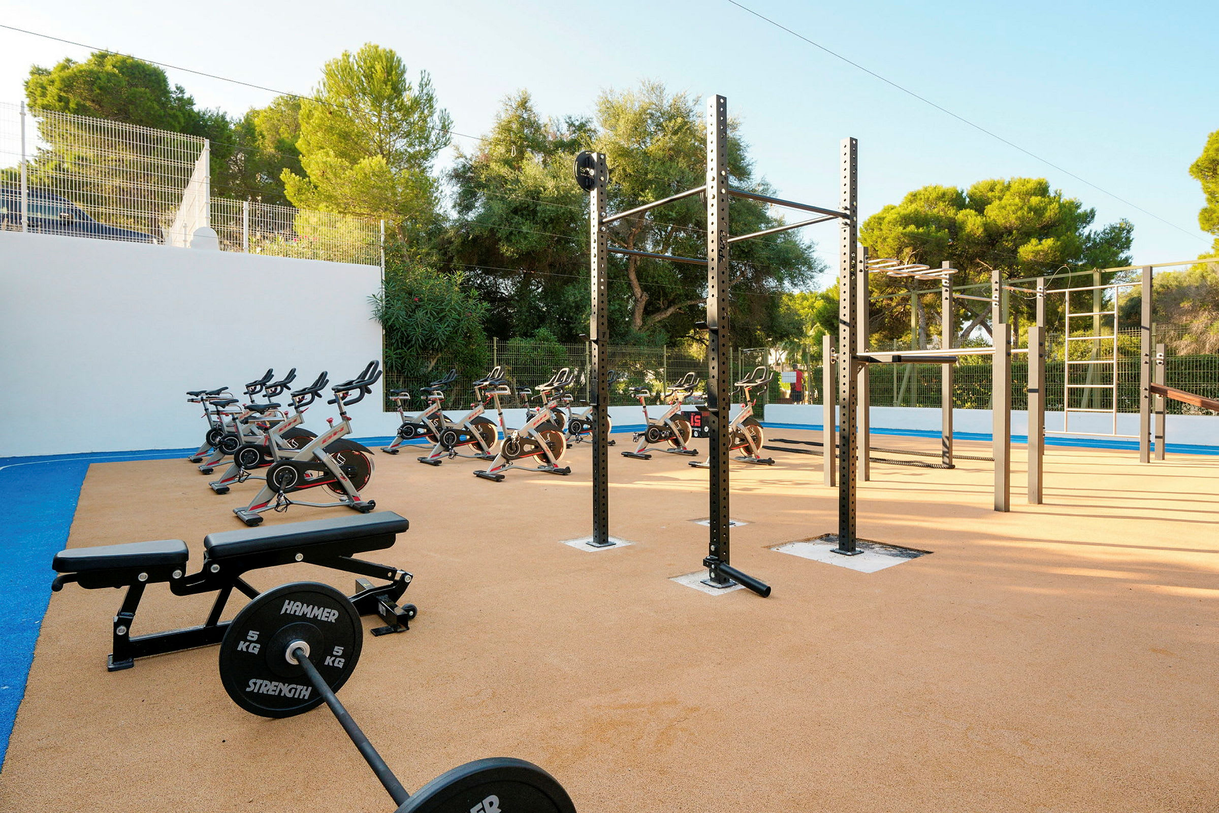 Utomhusgym med fitnessutrustning inklusive träningscyklar och vikter. Grönska och träd i bakgrunden. (AI-genererad text)