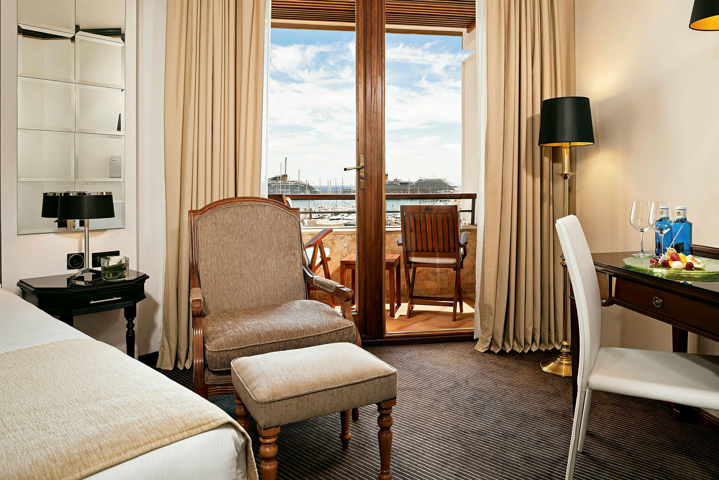 Ett hotellrum med en fåtölj, ett skrivbord och utsikt över en balkong med sittplats samt en hamn i bakgrunden. (AI-genererad text)