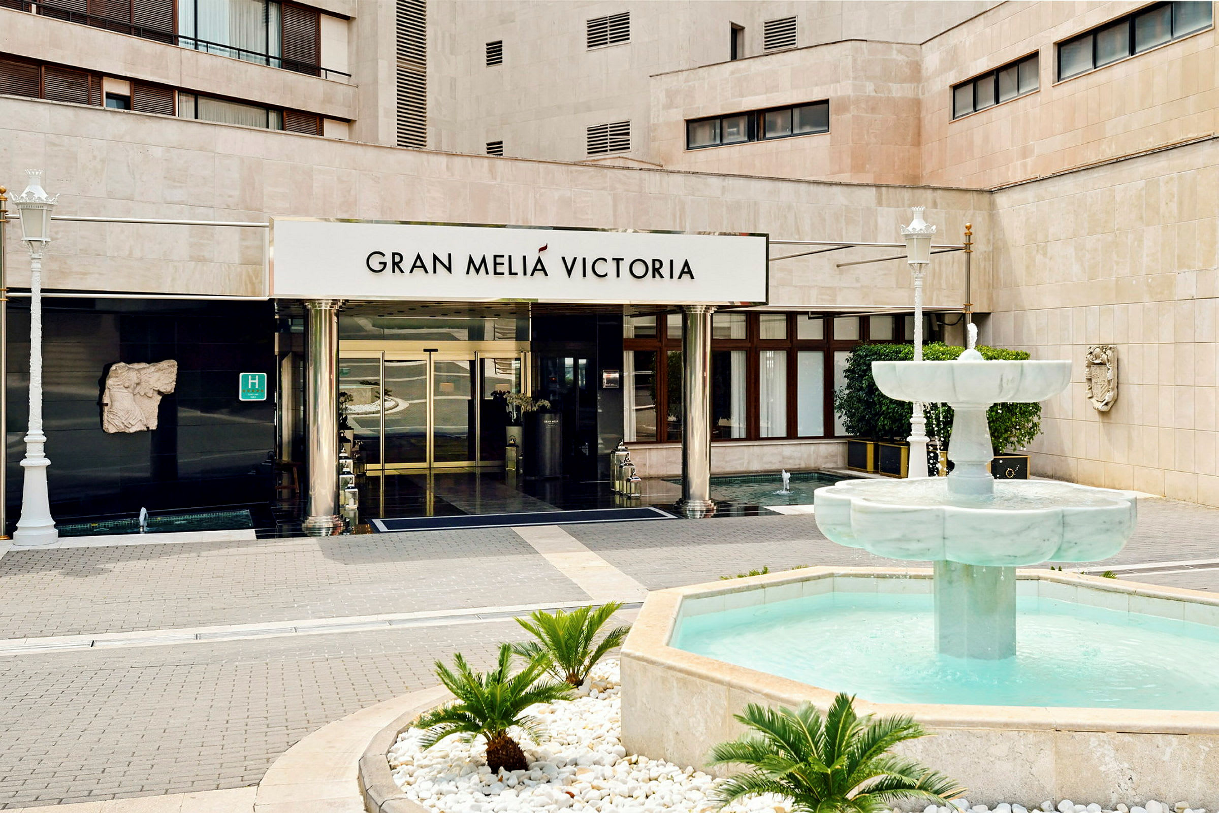 Entrén till en byggnad med texten "Gran Meliá Victoria" ovanför dörrarna samt en fontän med klart vatten framför. (AI-genererad text)