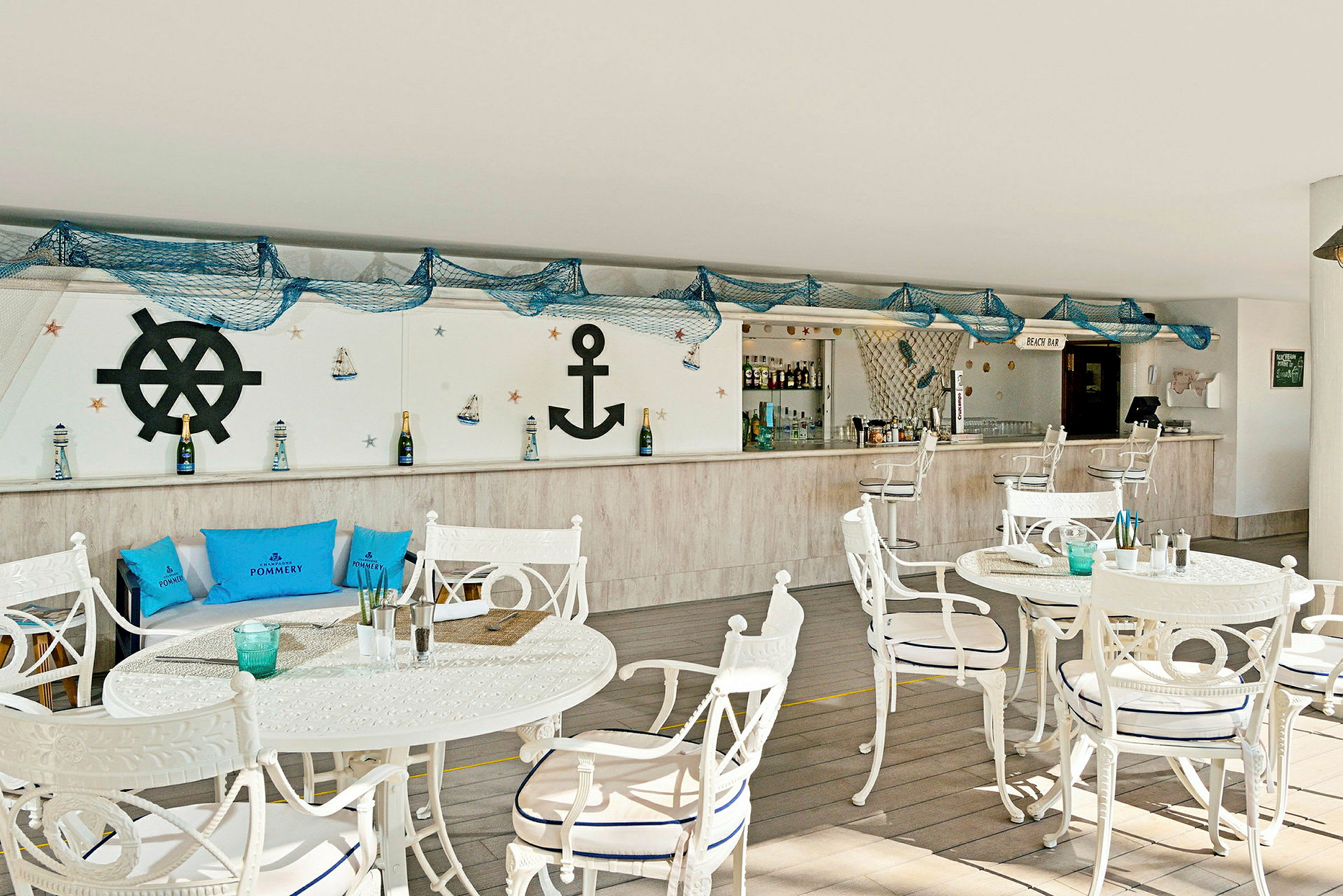 Bild av en strandbar med maritim inredning, inklusive vita bord och stolar, blåa kuddar och dekorationer som ankare och fiskenät. (AI-genererad text)