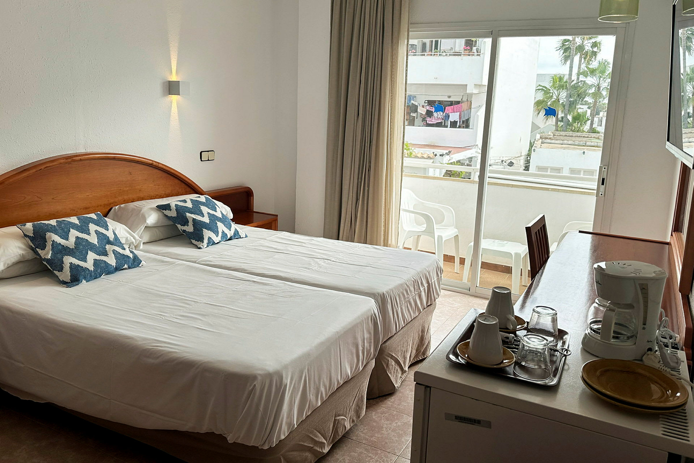 En hotellrum med två enkelsängar, ett skrivbord med kaffekokare och koppar, samt en balkong i bakgrunden. (AI-genererad text)