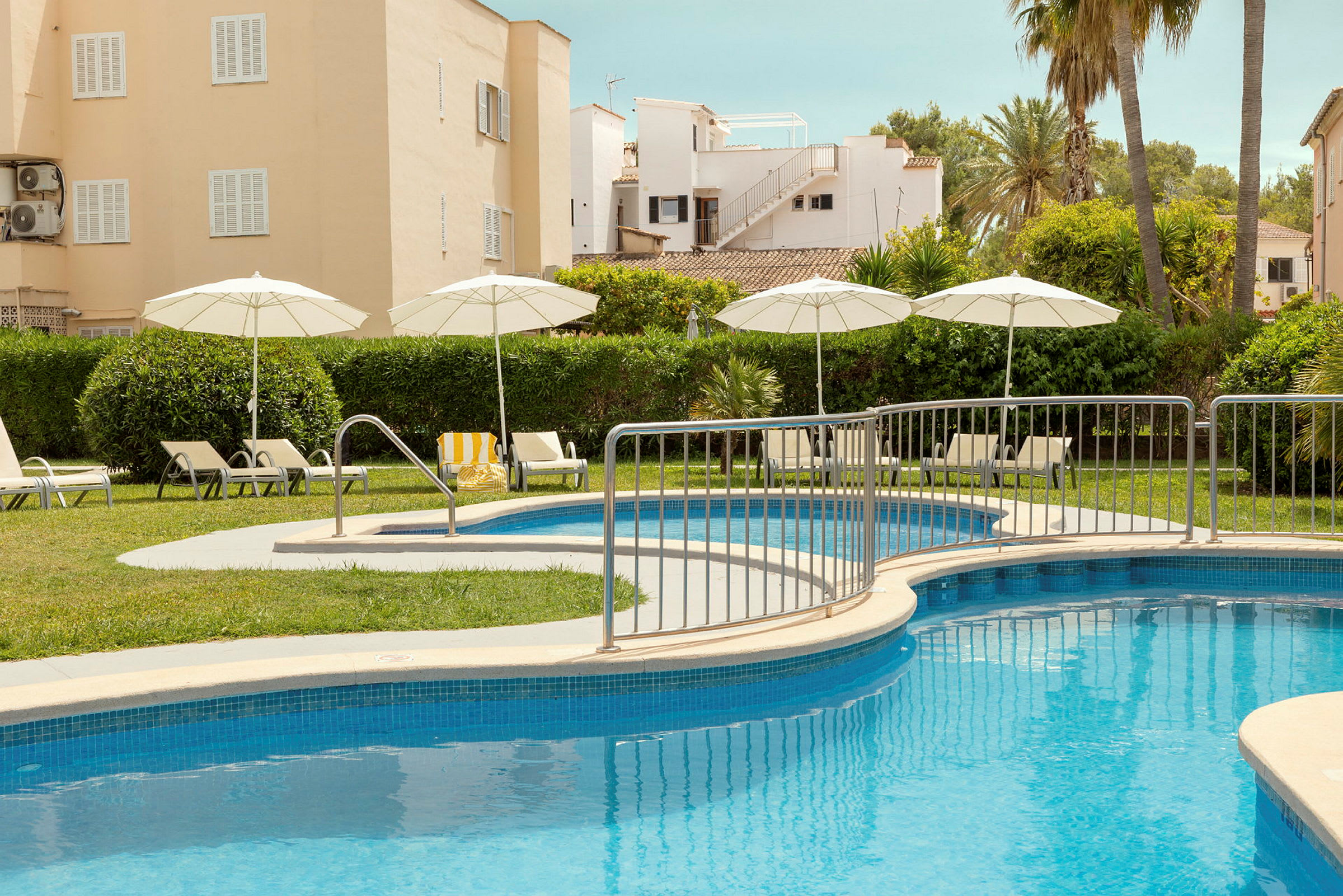 En pool med parasoller och solstolar framför ett bostadshus och grönska. (AI-genererad text)