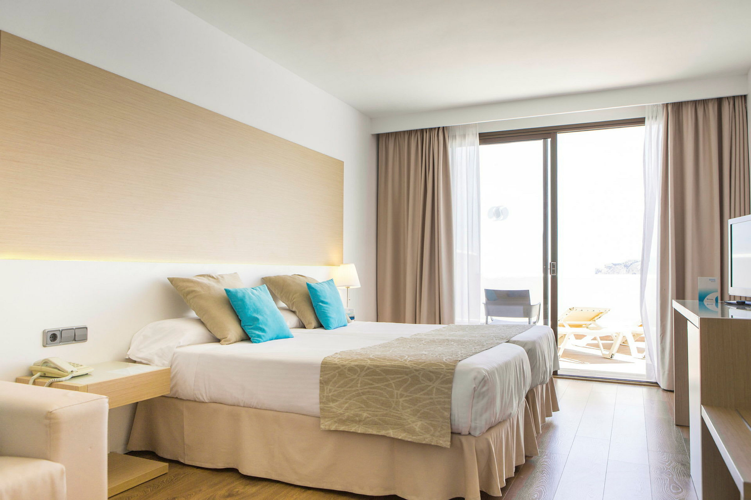 Ett hotellrum med en dubbelsäng, några kuddar, en balkongdörr och en TV. (AI-genererad text)