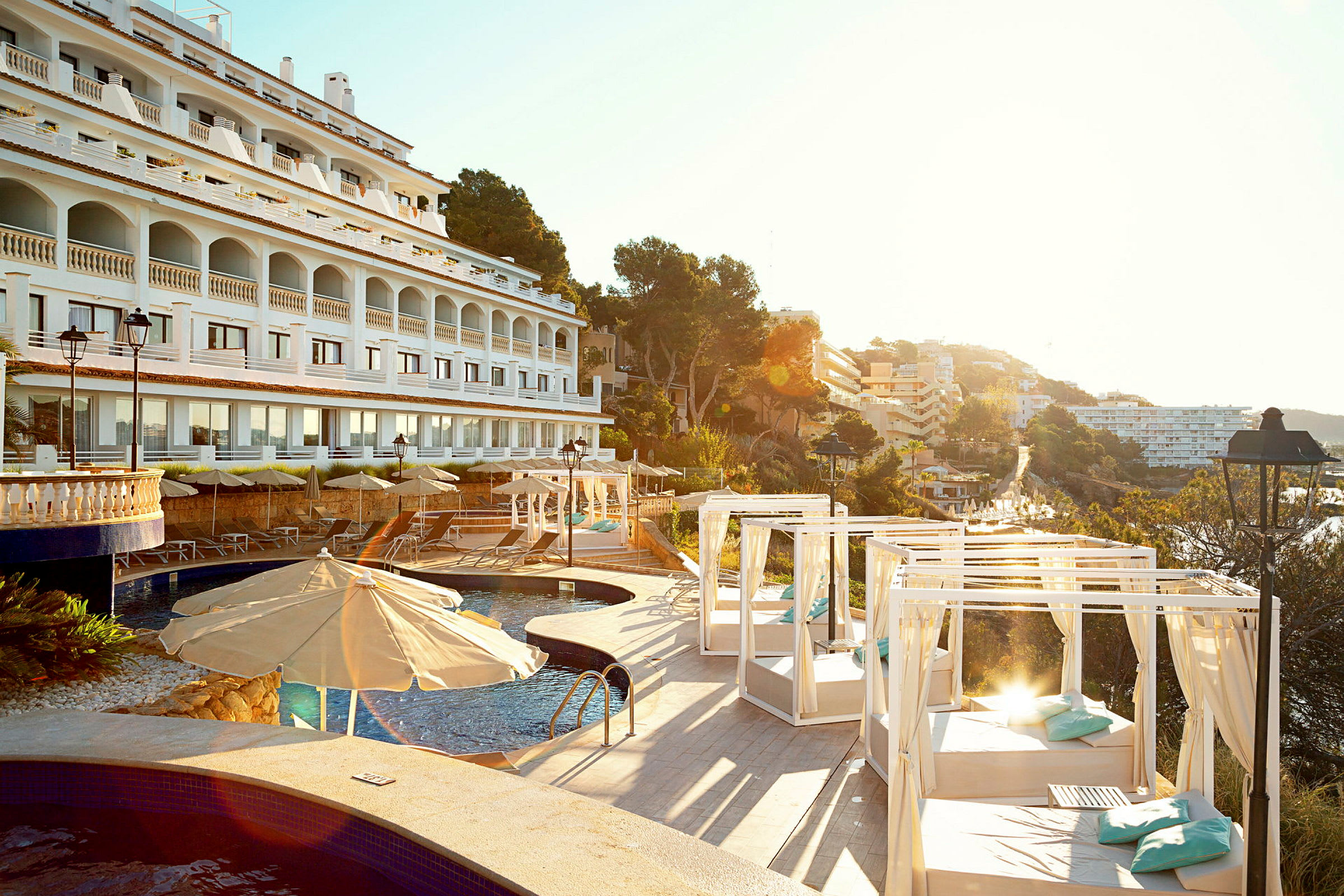 Hotellfasad i vit med arkader, swimmingpool och solstolar med parasoller och dagsängar, omgiven av träd och byggnader. (AI-genererad text)