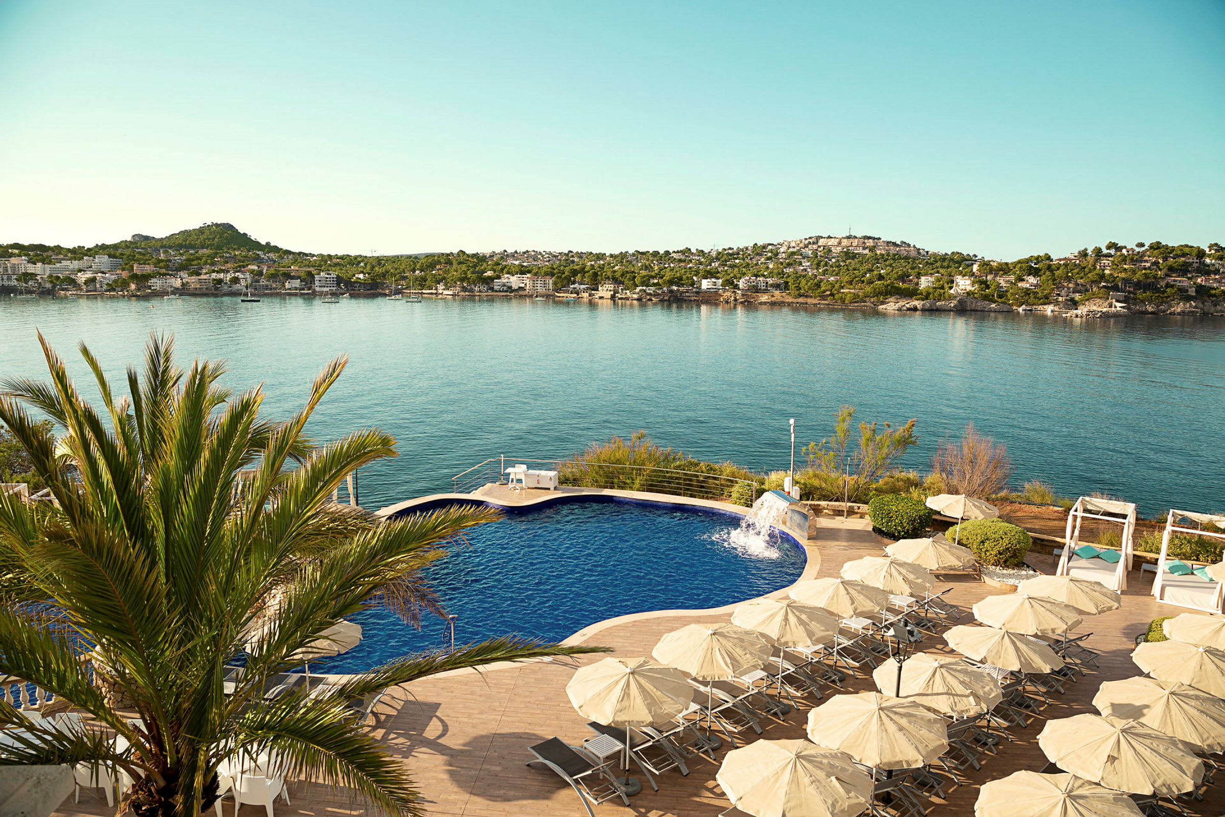 En pool med solstolar och parasoller. I bakgrunden syns havet och en kustlinje med hus. (AI-genererad text)
