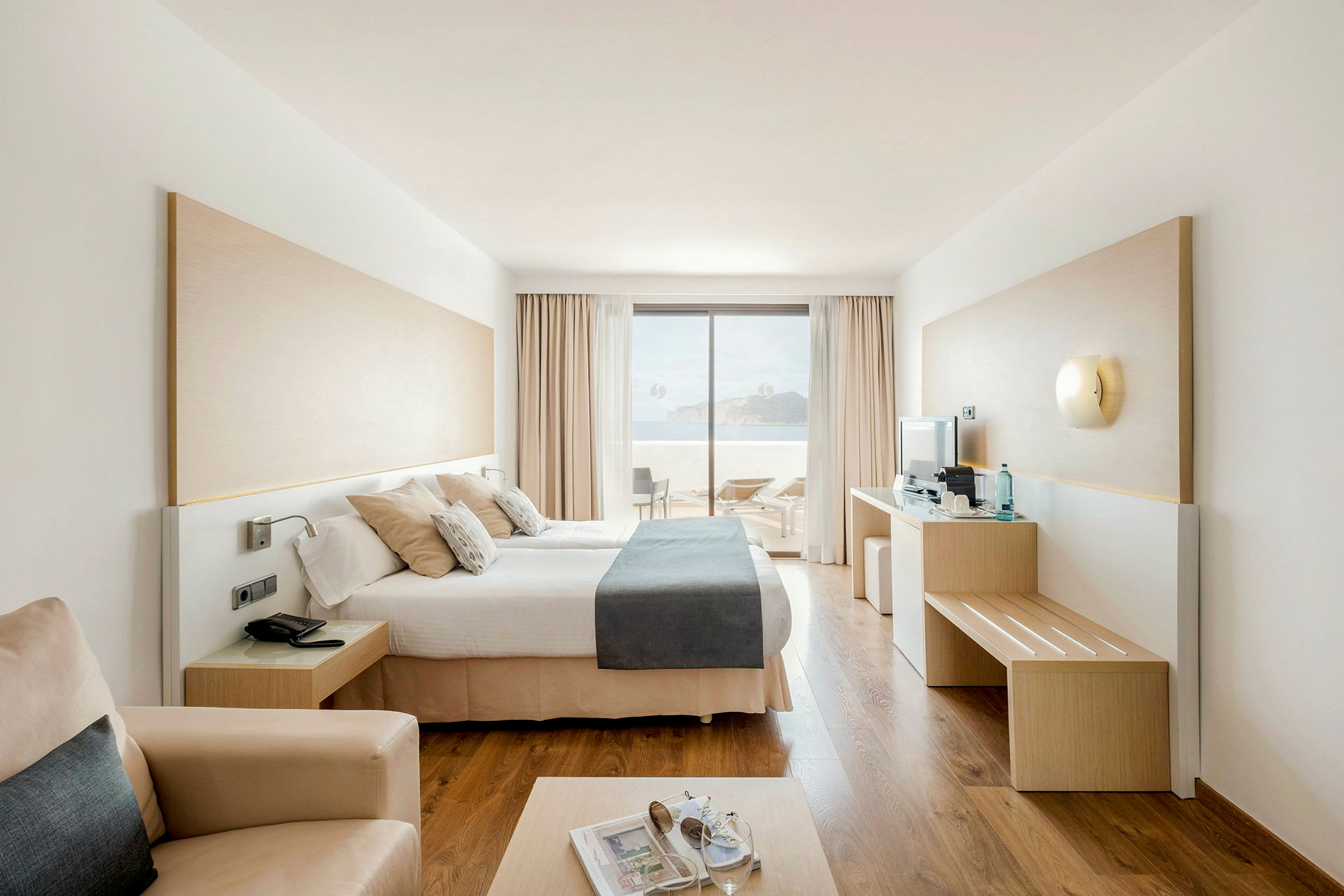 Modernt hotellrum med dubbelsäng och utsikt mot terrass och hav. (AI-genererad text)