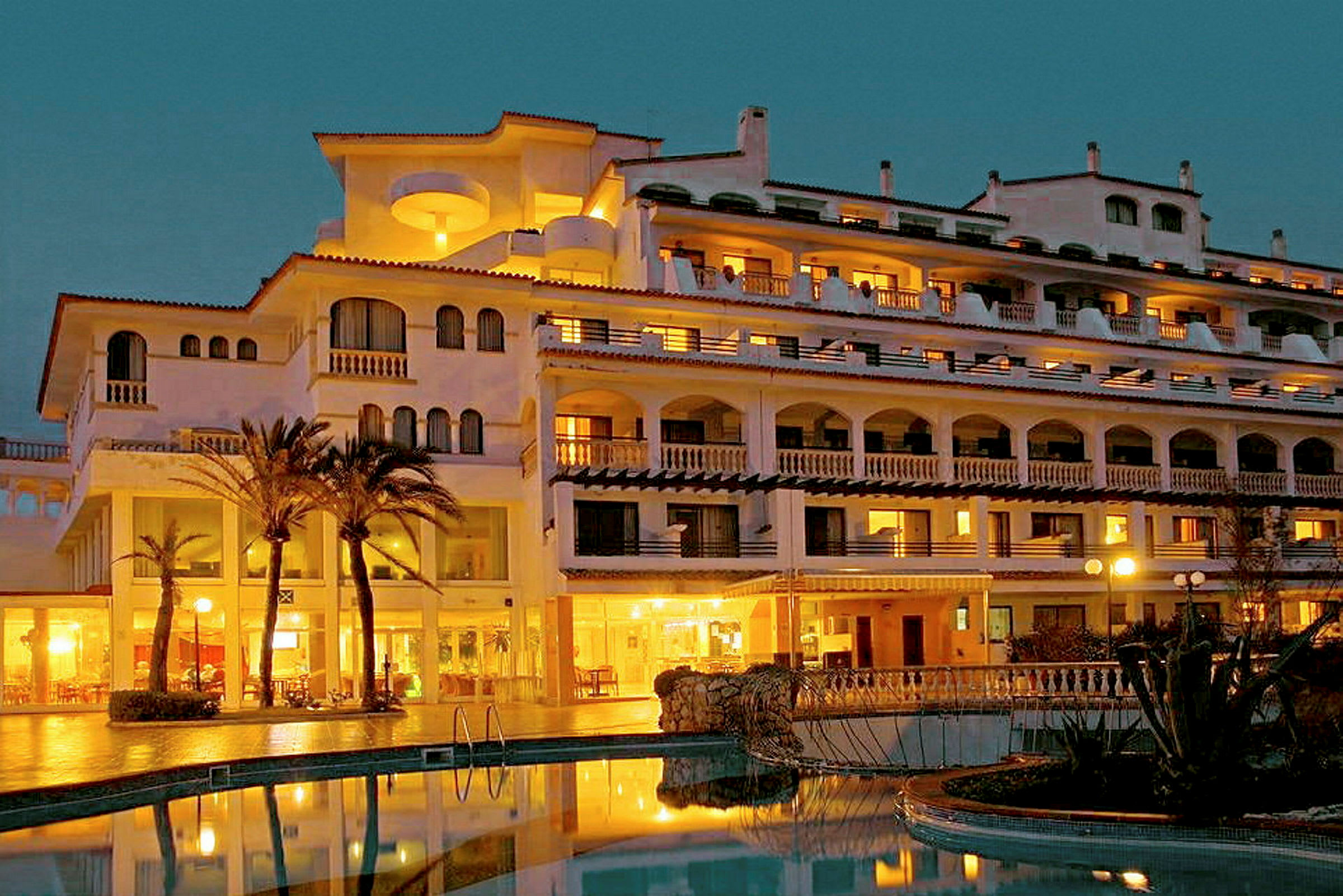 Ett fler våningshotell med upplysta balkonger och palmer framför en pool. (AI-genererad text)