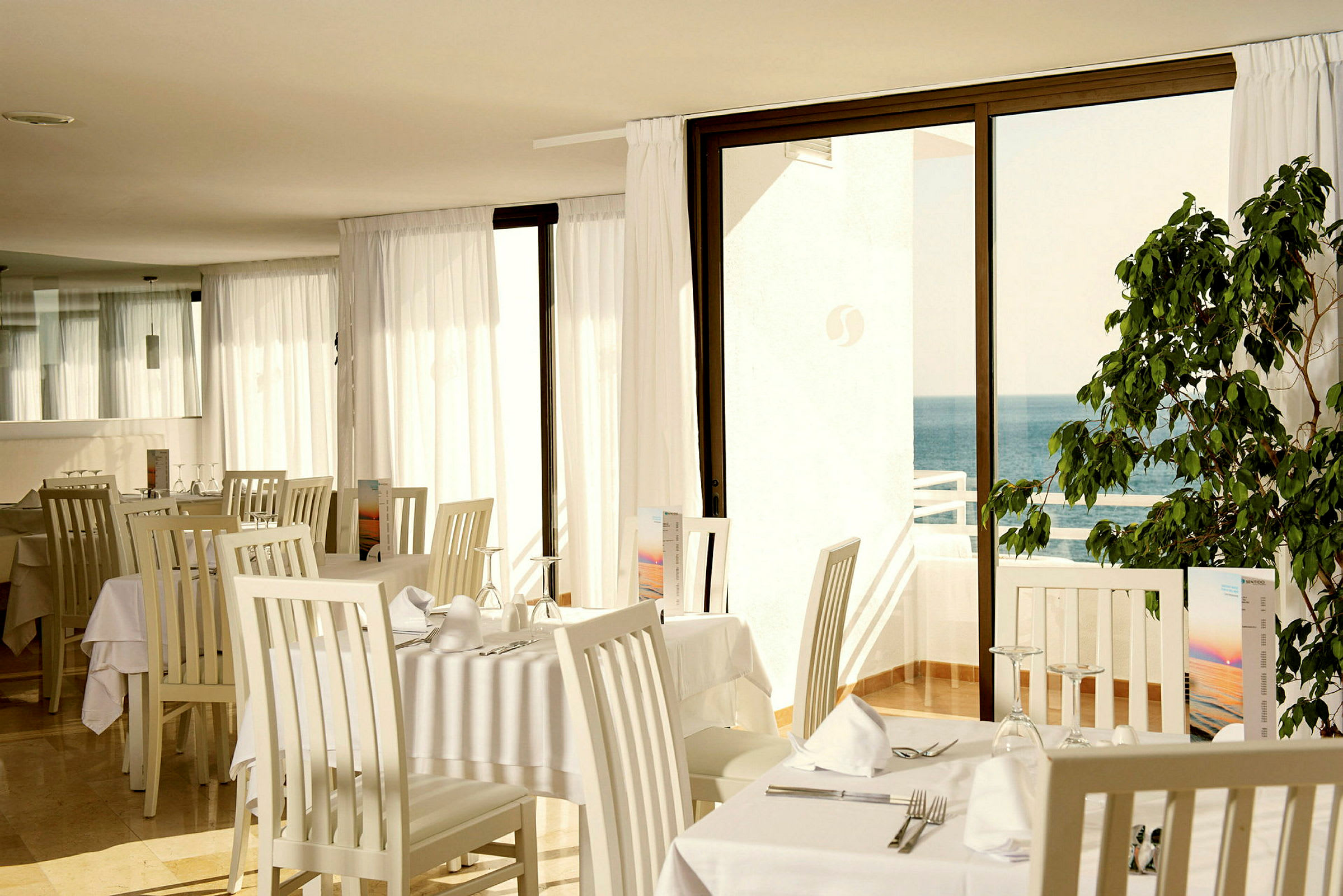 En restaurang med vita borddukade bord, ljusa stolar och glasdörrar som öppnar upp mot havet. (AI-genererad text)